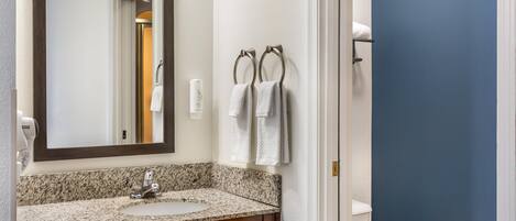 A bathroom with a granite countertop sink, a mirror, a towel rack with two towels, and a toilet.