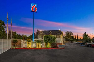 Un motel con un llamativo letrero con el número '6', un camino de entrada con portones y un estacionamiento con varios vehículos.