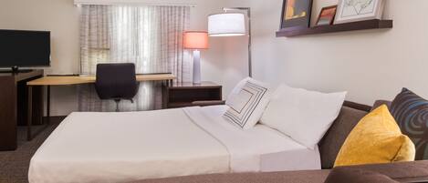 A hotel room with a bed, desk, chair, TV, lamp, and artwork on the wall.