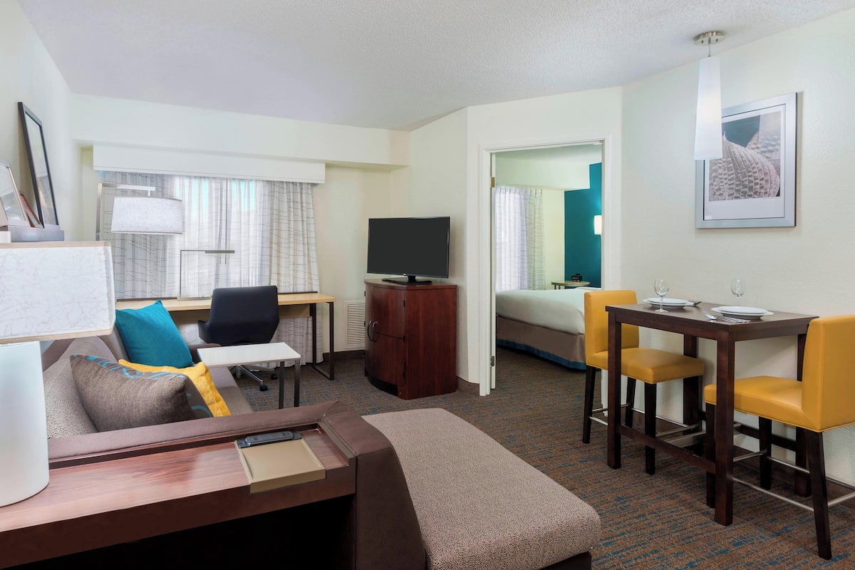 A hotel room with a bed, a desk, a television, a dining table with chairs, and a window with curtains.