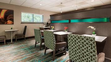 A hotel room with a patterned carpet, a dining area with chairs and tables, a window with a view, and a wall with a green accent panel.