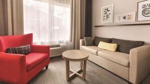 Una sala de estar con un sillón rojo, un sofá color beige, una mesita de madera y un ventanal con cortinas traslúcidas.