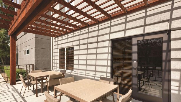 Un patio exterior con una pérgola de madera, una mesa y sillas, y un ventanal corredero que da a un área de patio.