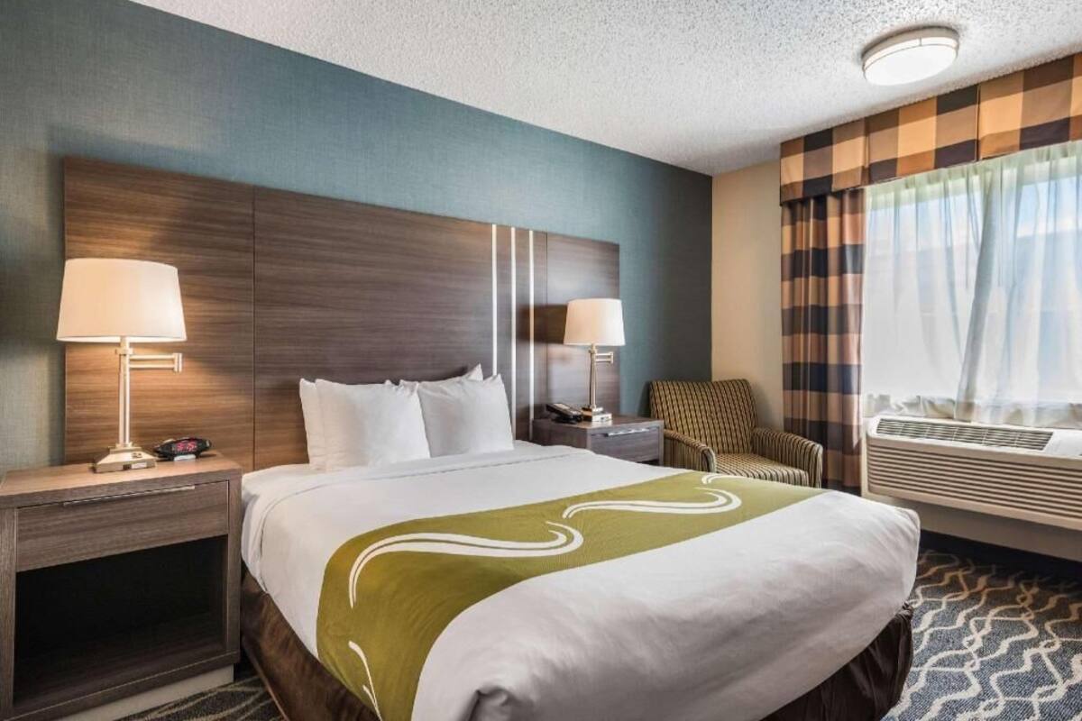 A hotel room with a large bed, two bedside tables with lamps, a chair, an air conditioning unit, and a window with curtains.