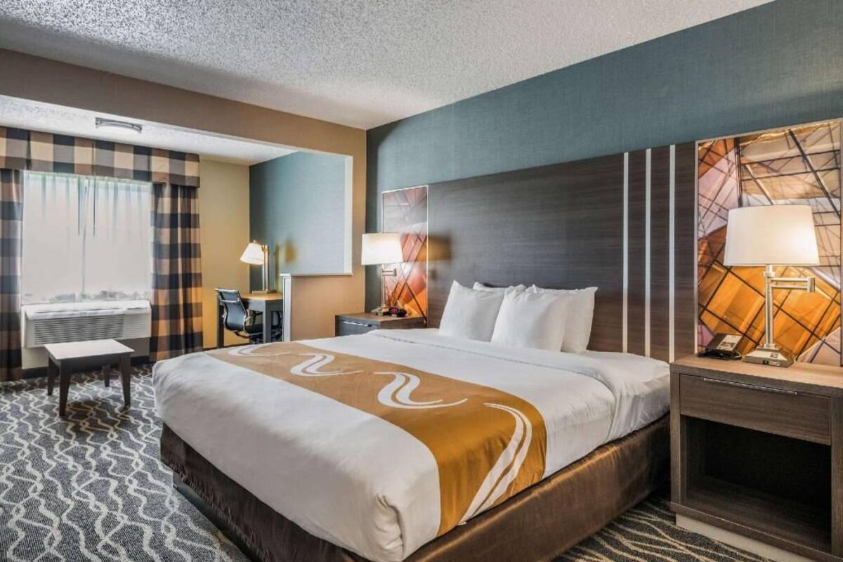 A hotel room with a large bed, a desk, a chair, a lamp, and a window with curtains.