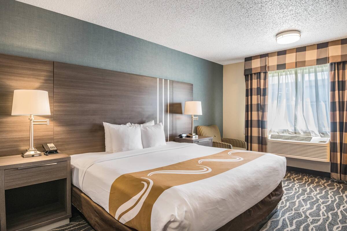 A hotel room with a large bed, two bedside tables with lamps, a chair, a window with curtains, and a ceiling light.