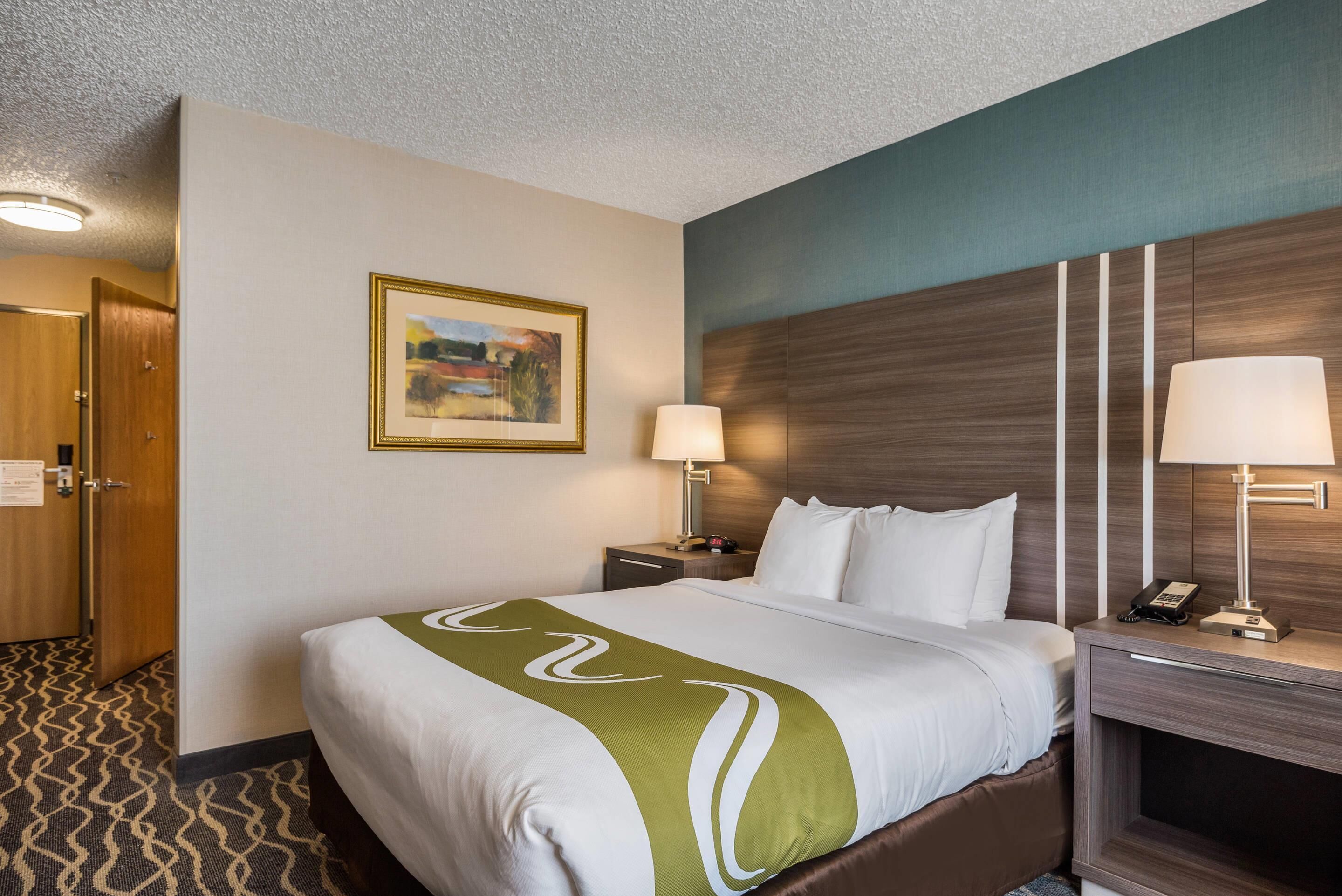 A hotel room with a neatly made bed, a nightstand with a lamp, a framed picture on the wall, and a door to the left.
