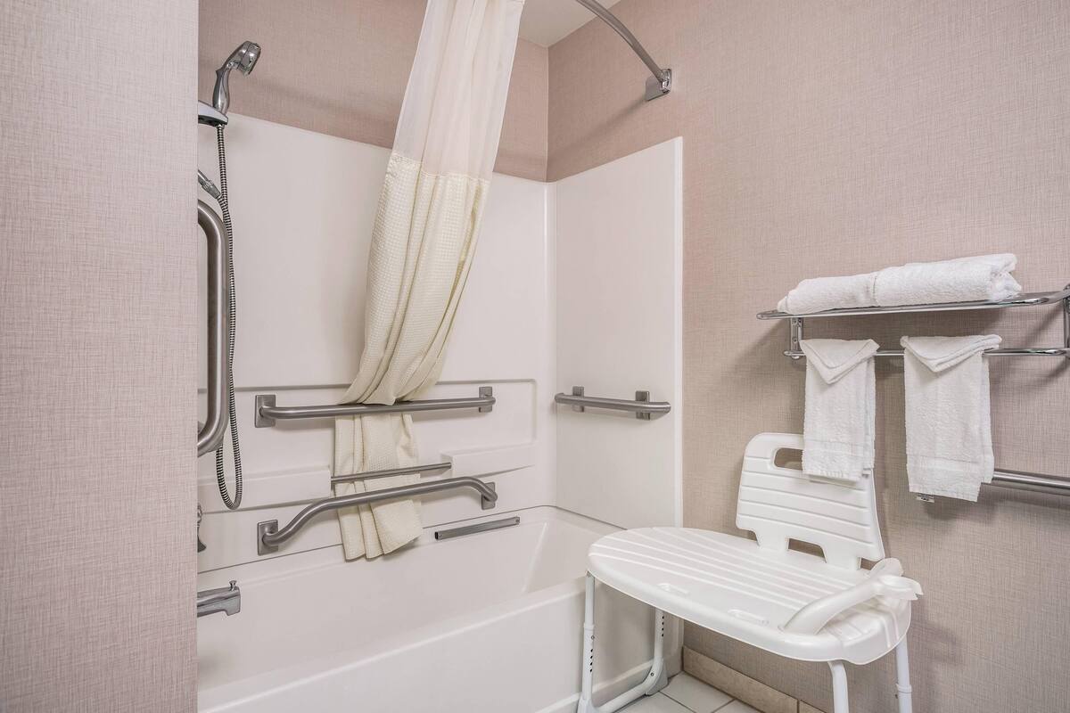 A bathroom with a shower, grab bars, a white chair, and a towel rack.