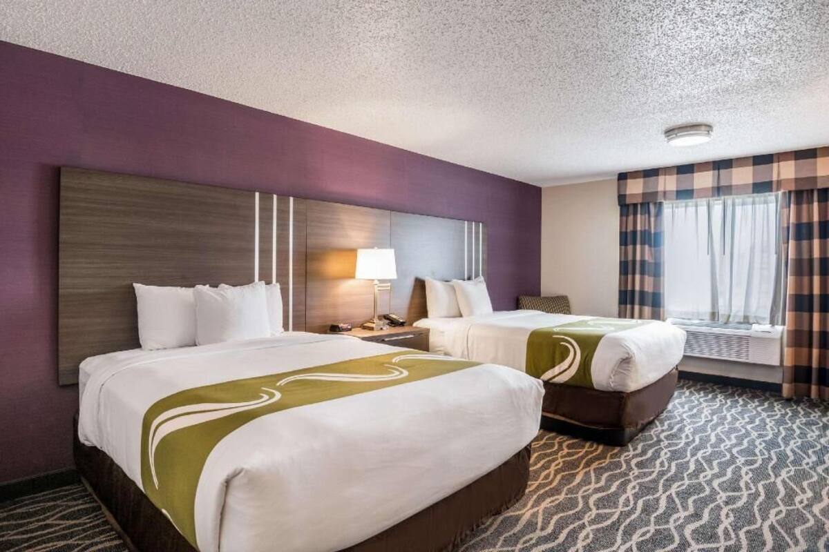 A hotel room with two beds, a large headboard, a nightstand, a lamp, a window with curtains, and a patterned carpet.