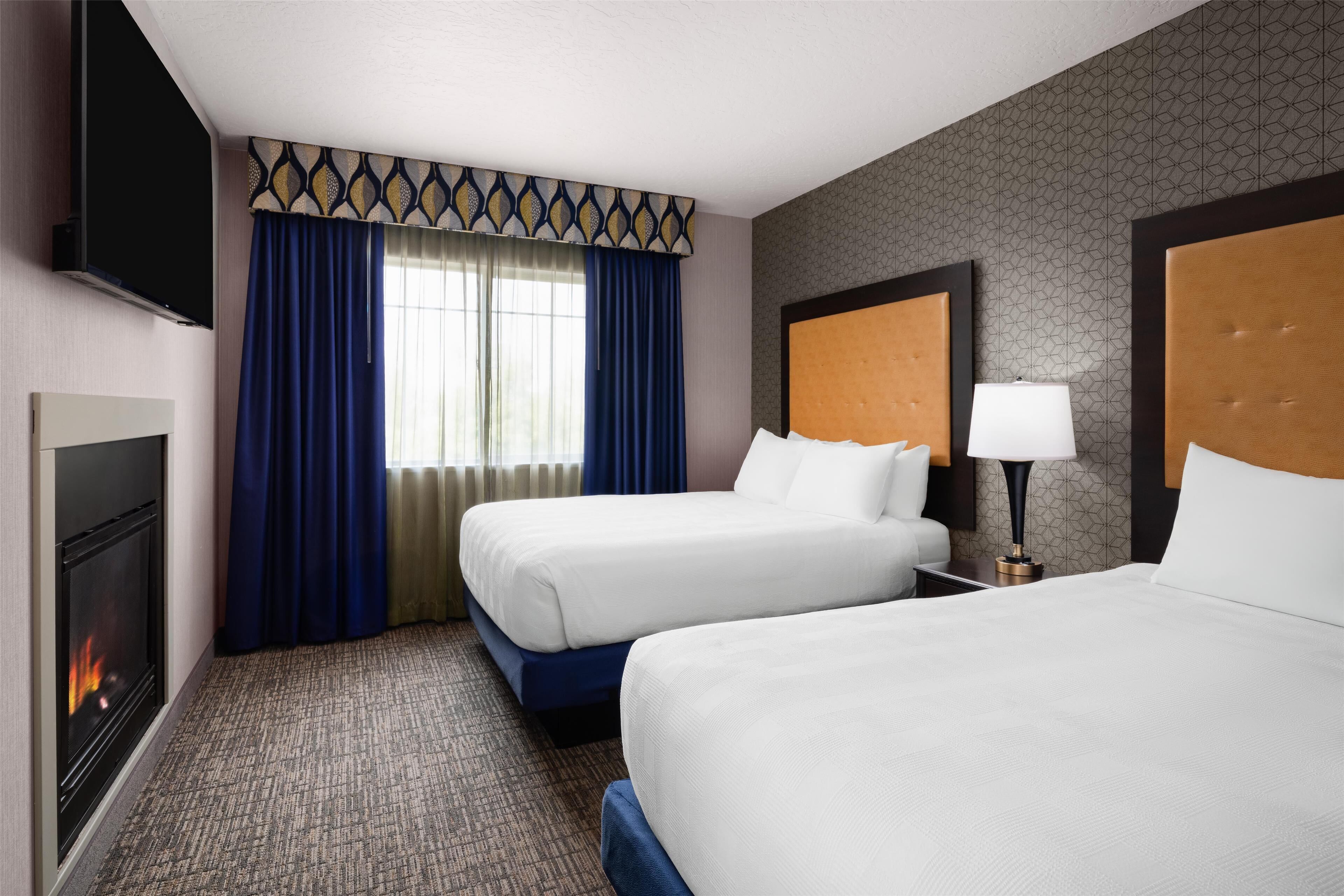 A hotel room with two beds, a TV, a fireplace, and a window with curtains.