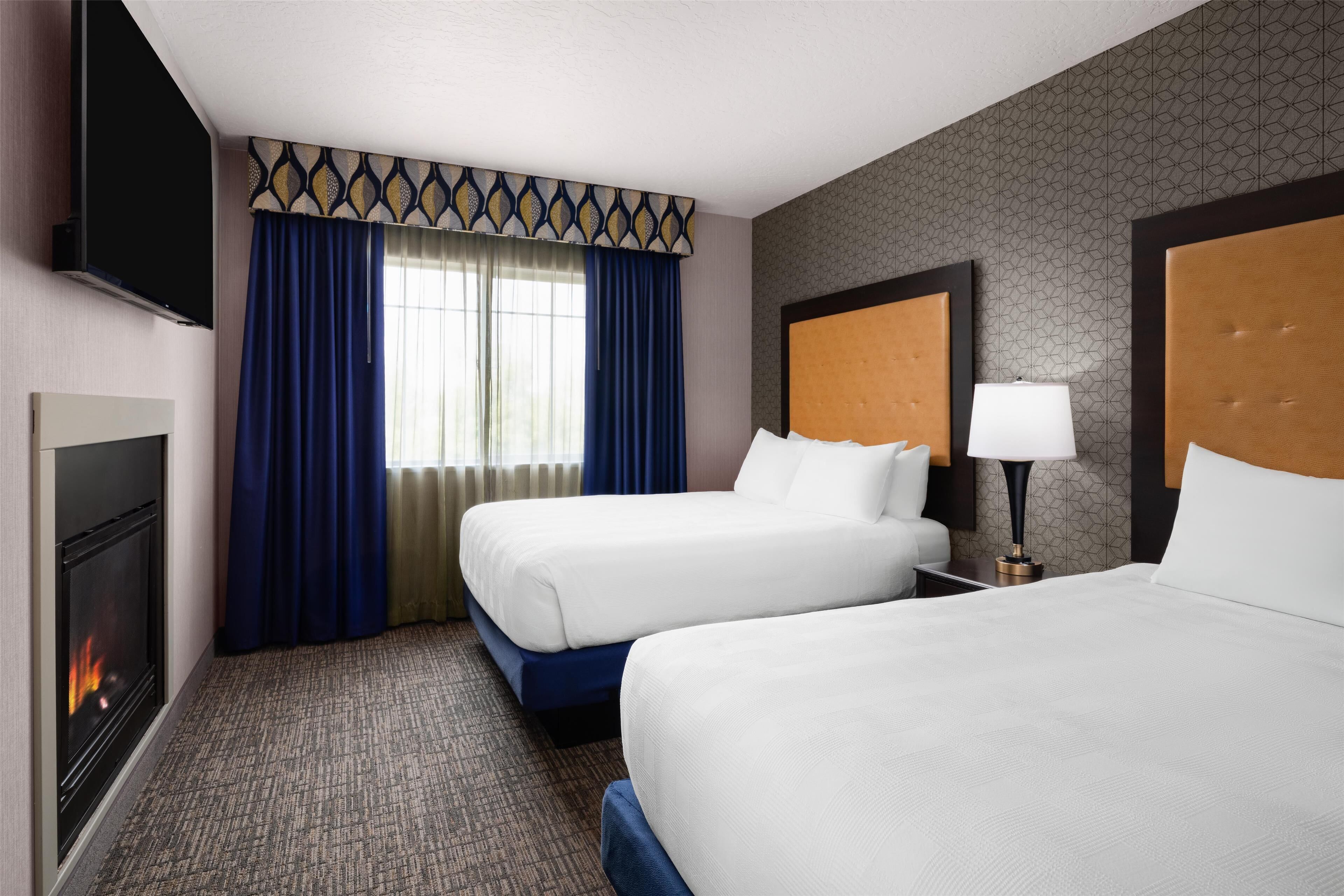 A hotel room with two beds, a television, a fireplace, and a window with curtains.