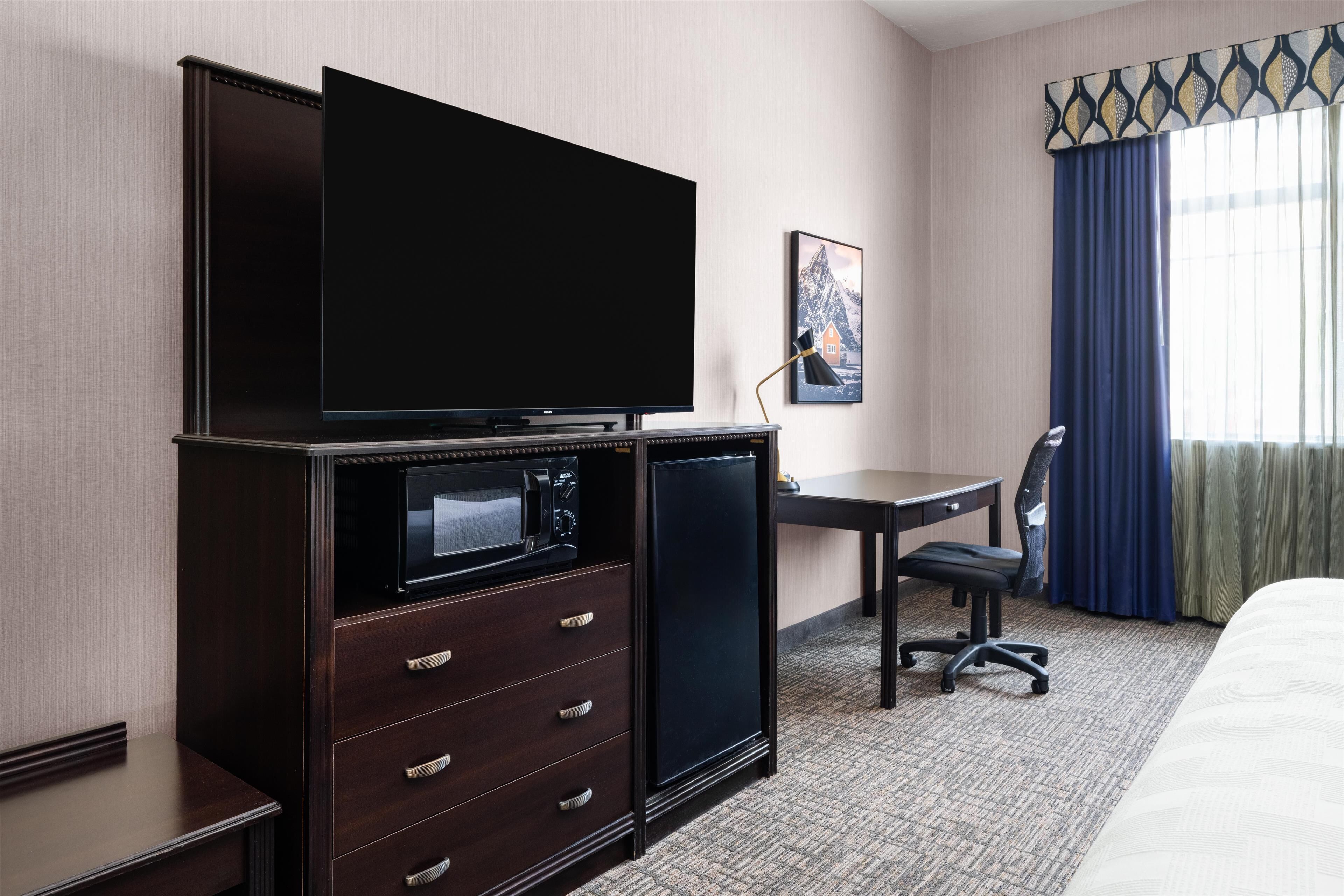 A hotel room with a flat-screen TV, a minibar, a desk with a chair, and a bed.