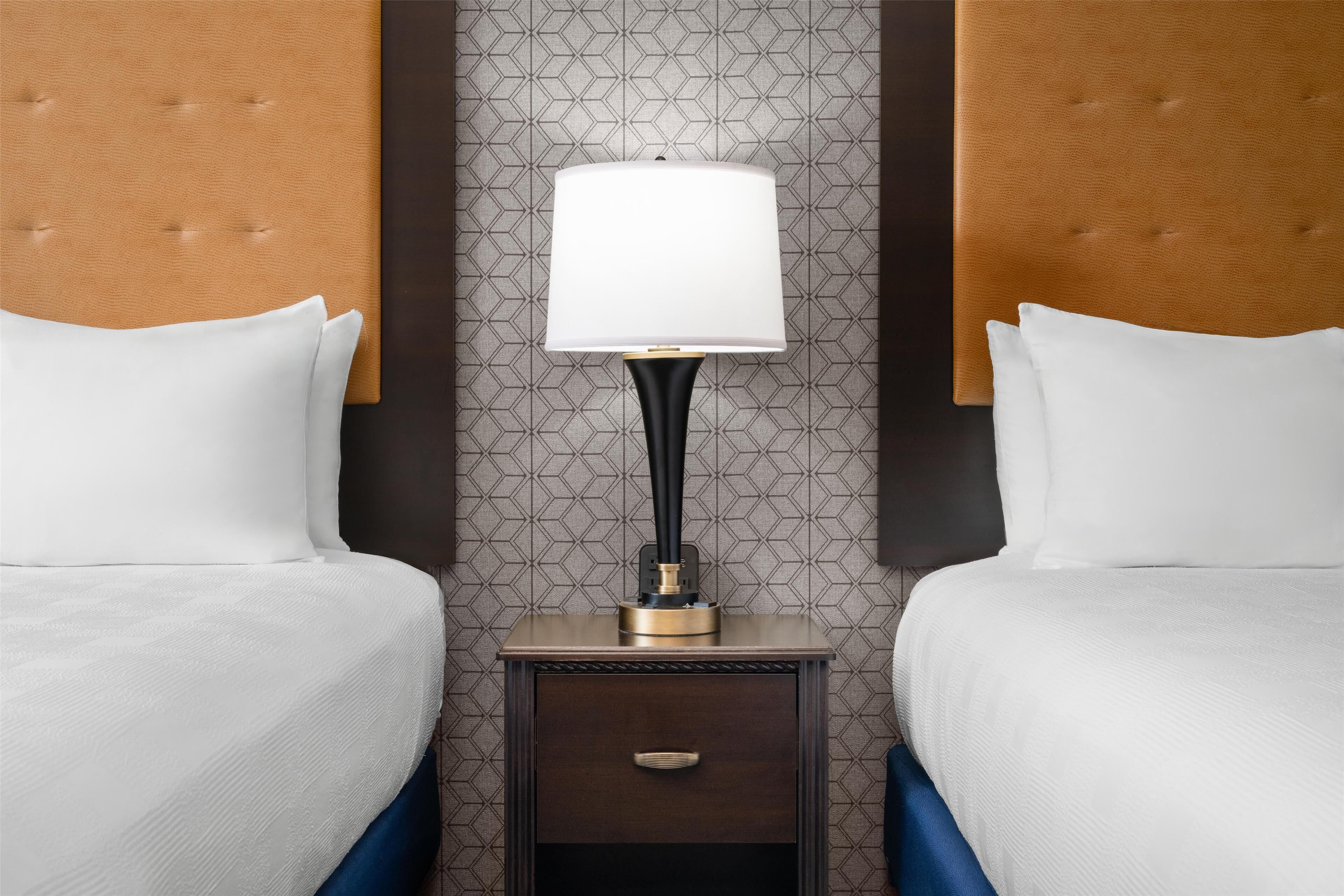 Quarto de hotel com duas camas, uma mesa de cabeceira com abajur e papel de parede estampado.
