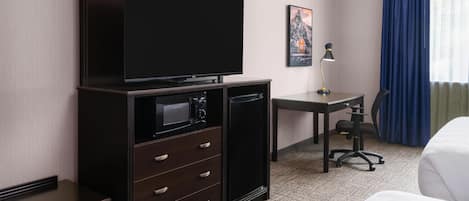 A hotel room with a flat-screen TV, a desk with a lamp, a chair, and a bed.
