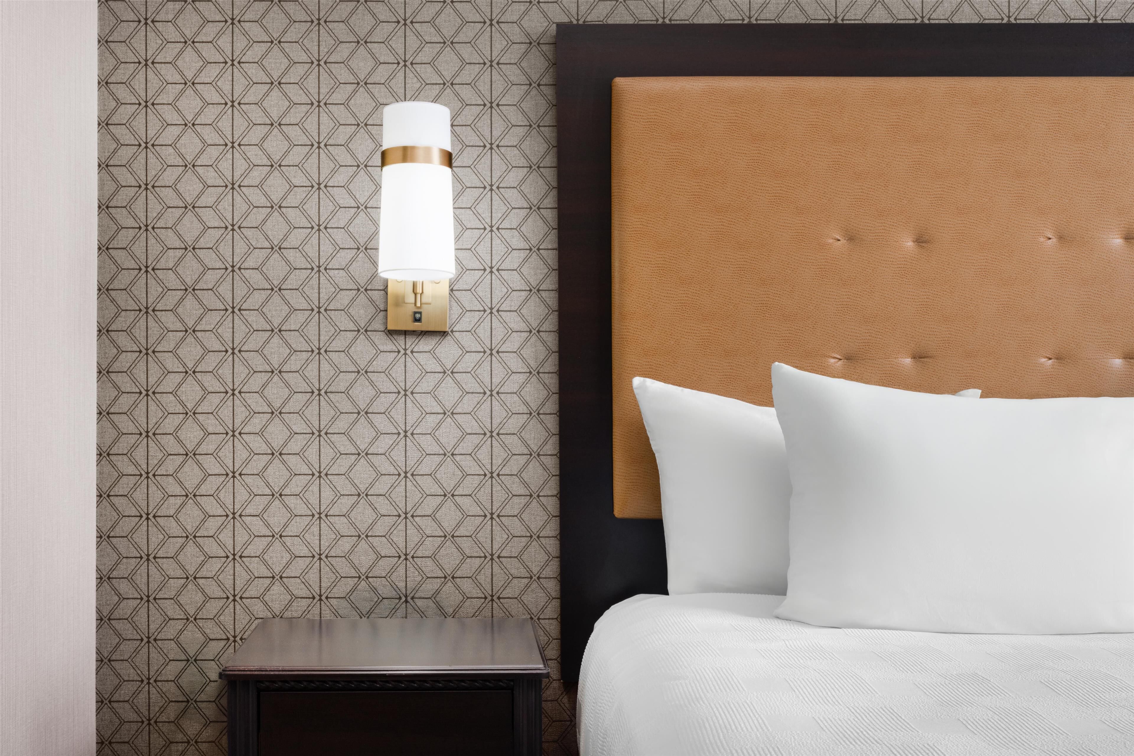 Une chambre d’hôtel avec un papier peint à motifs, un luminaire fixé au mur, une tête de lit capitonnée et un lit soigneusement fait avec du linge de lit blanc.
