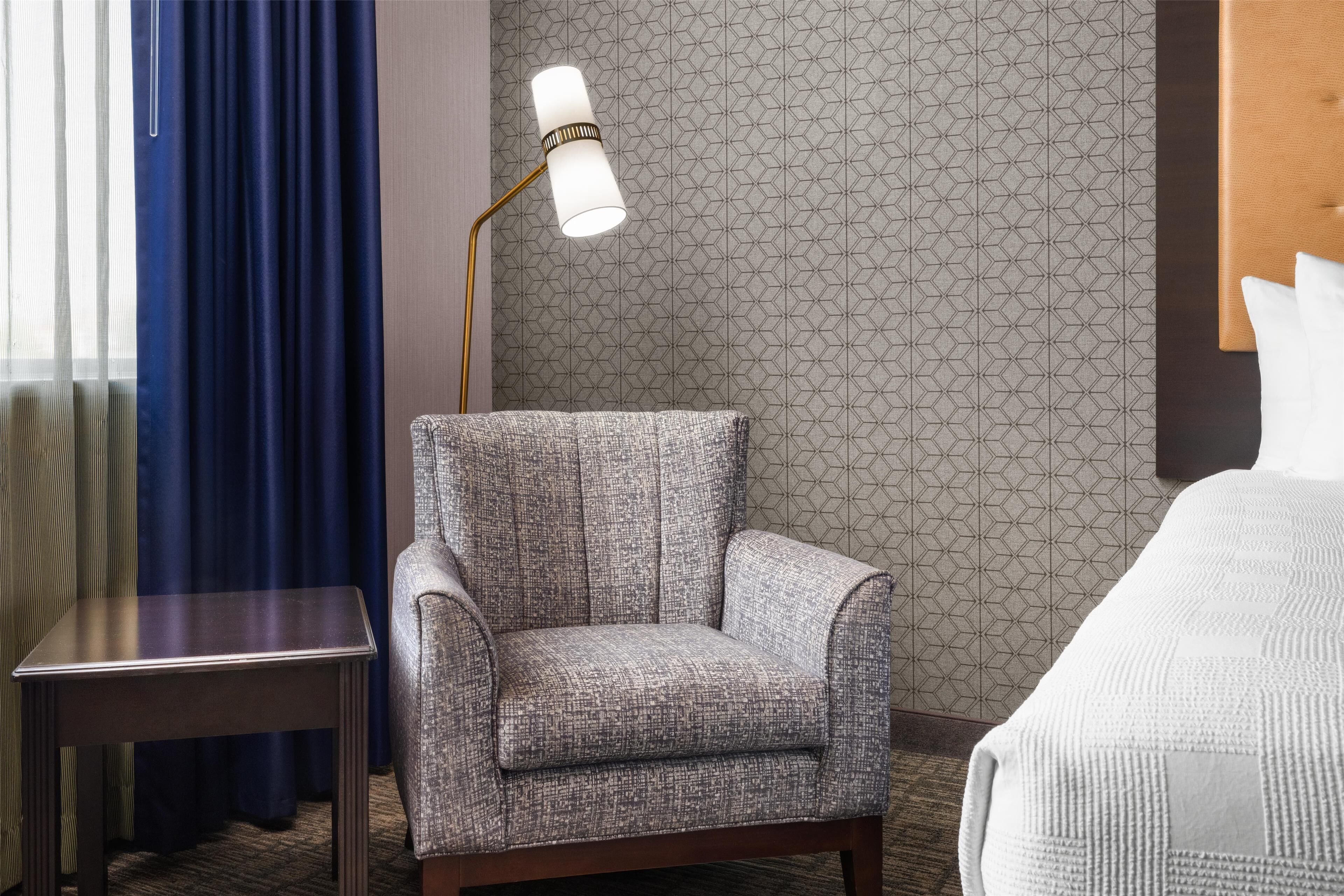 Une chambre d’hôtel avec un mur à motifs, un fauteuil, une table de chevet, une lampe et un lit avec des oreillers.