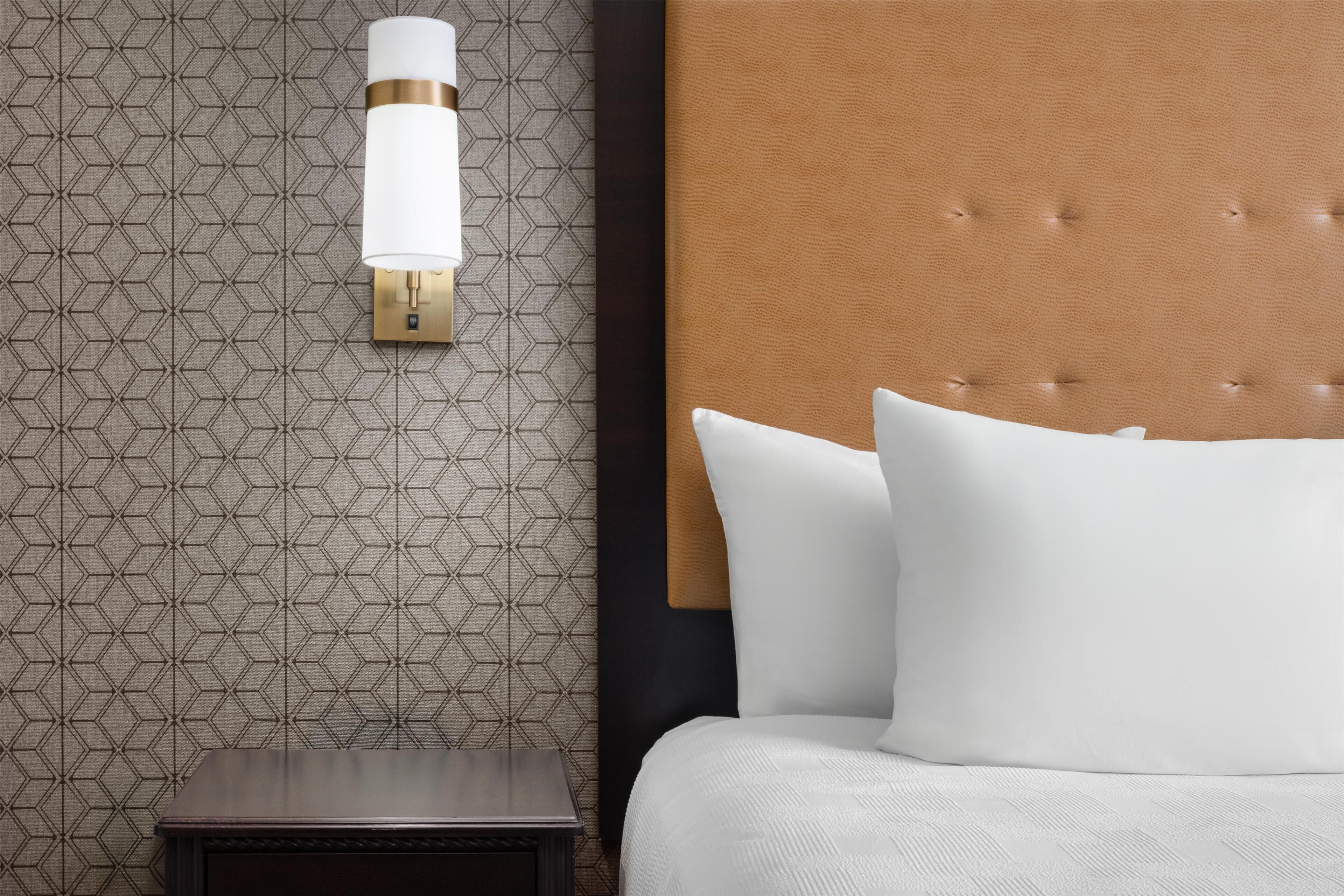 A hotel room with a patterned wallpaper, a bedside lamp, a wooden nightstand, and a bed with white bedding.