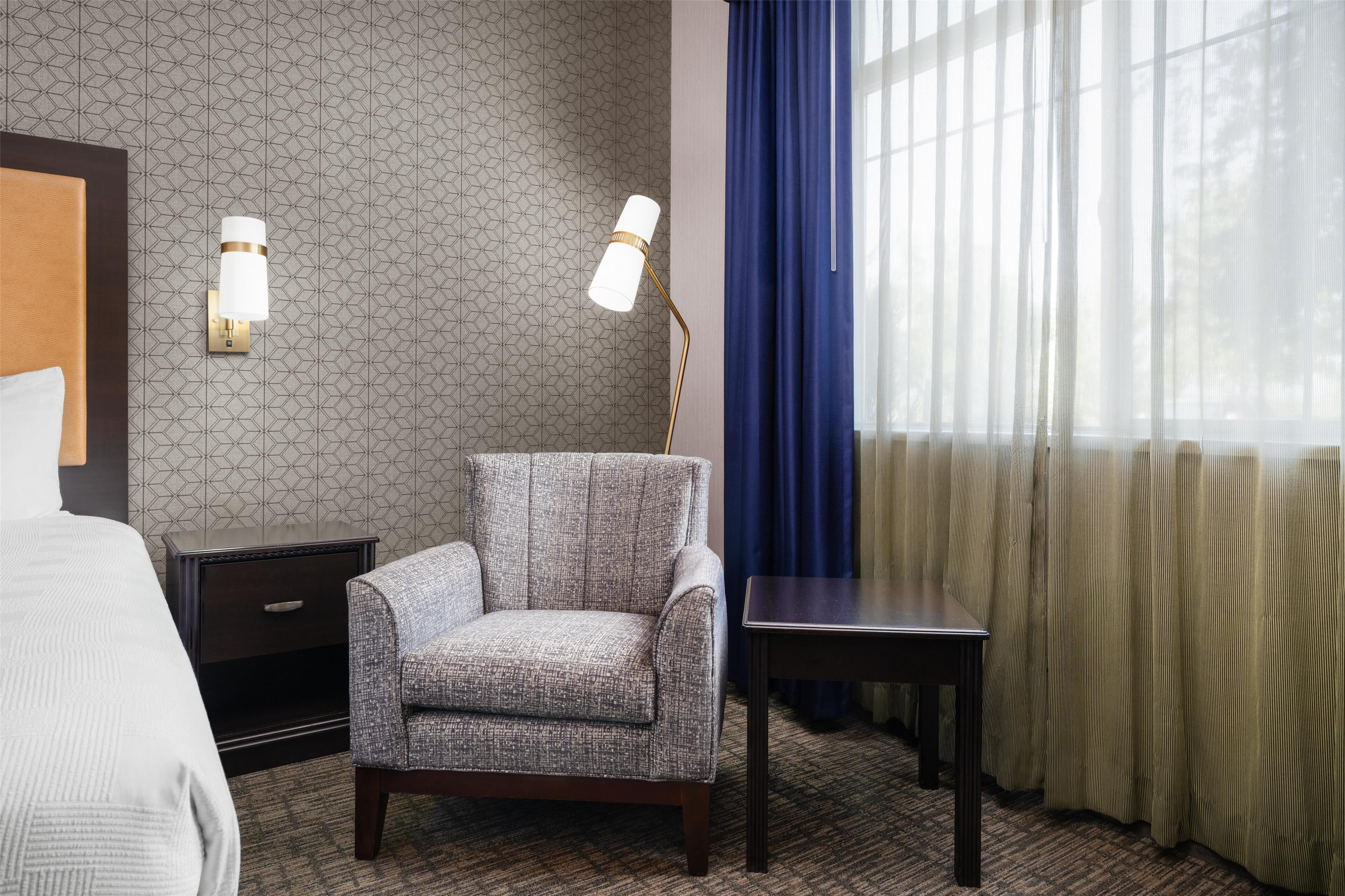Habitación de hotel con pared estampada, un sillón gris, una mesita, una mesita de noche y una cama.