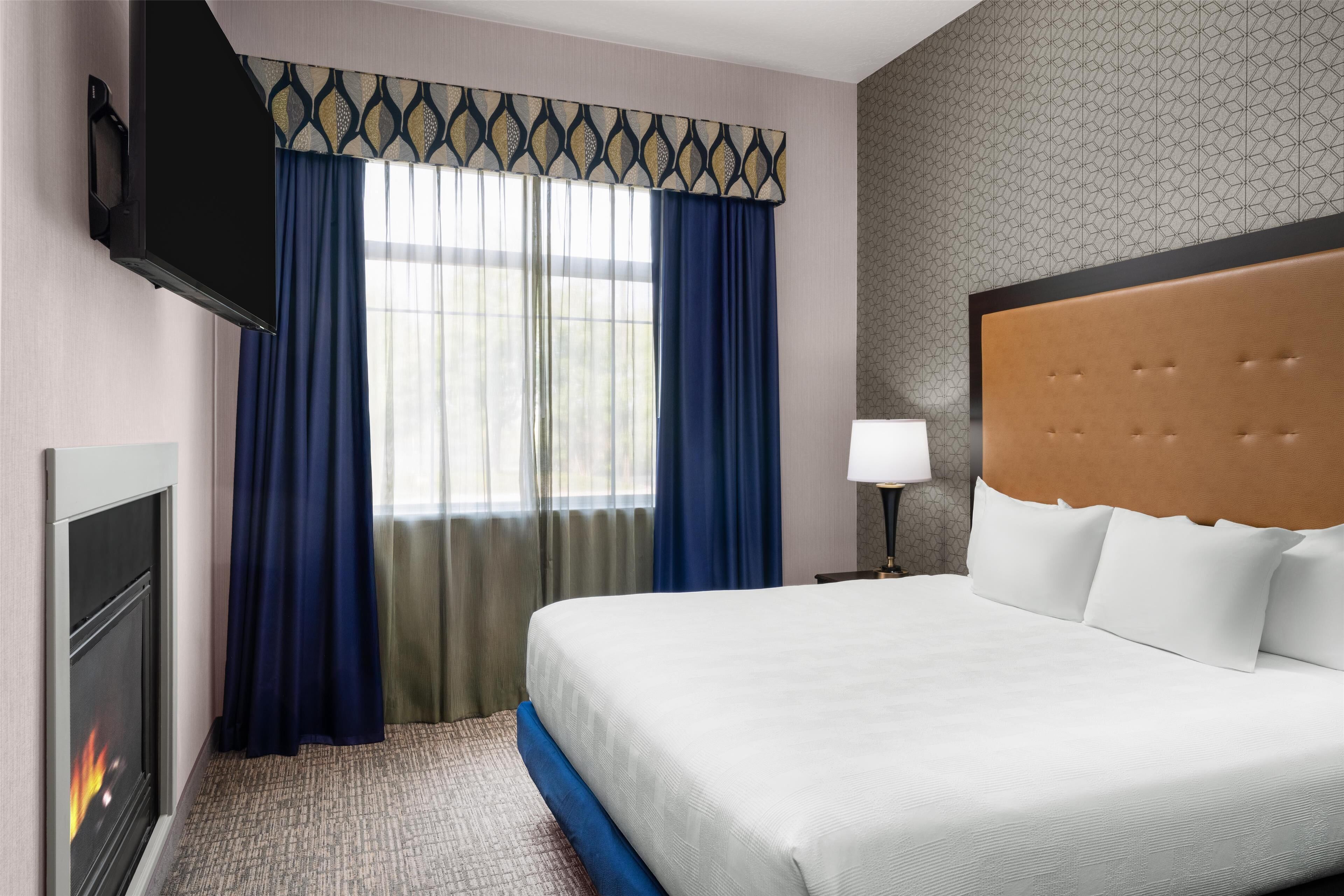 A hotel room with a bed, a TV, a fireplace, and a window with curtains.