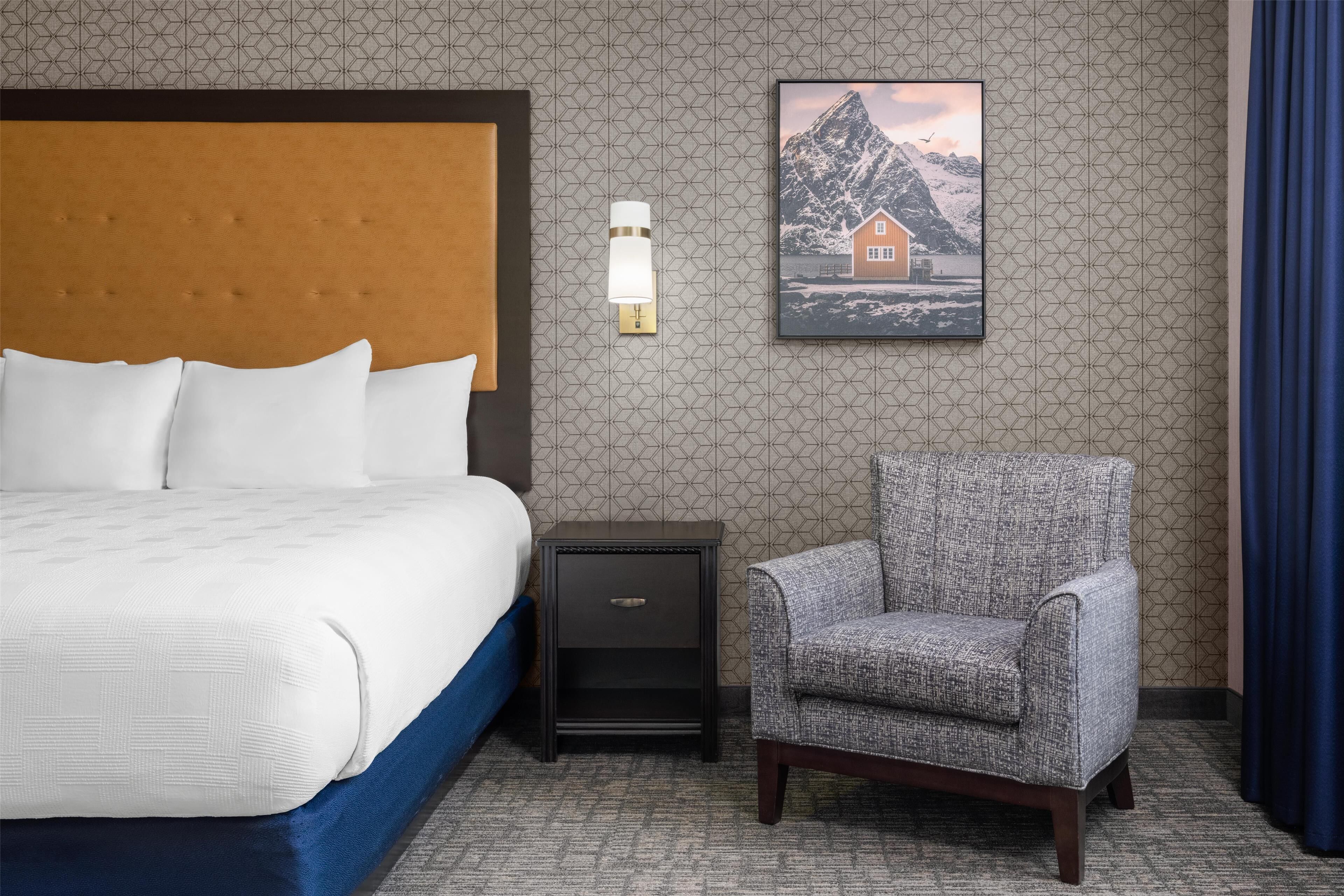 A hotel room with a bed, a nightstand, a chair, a wall-mounted lamp, and a framed picture.