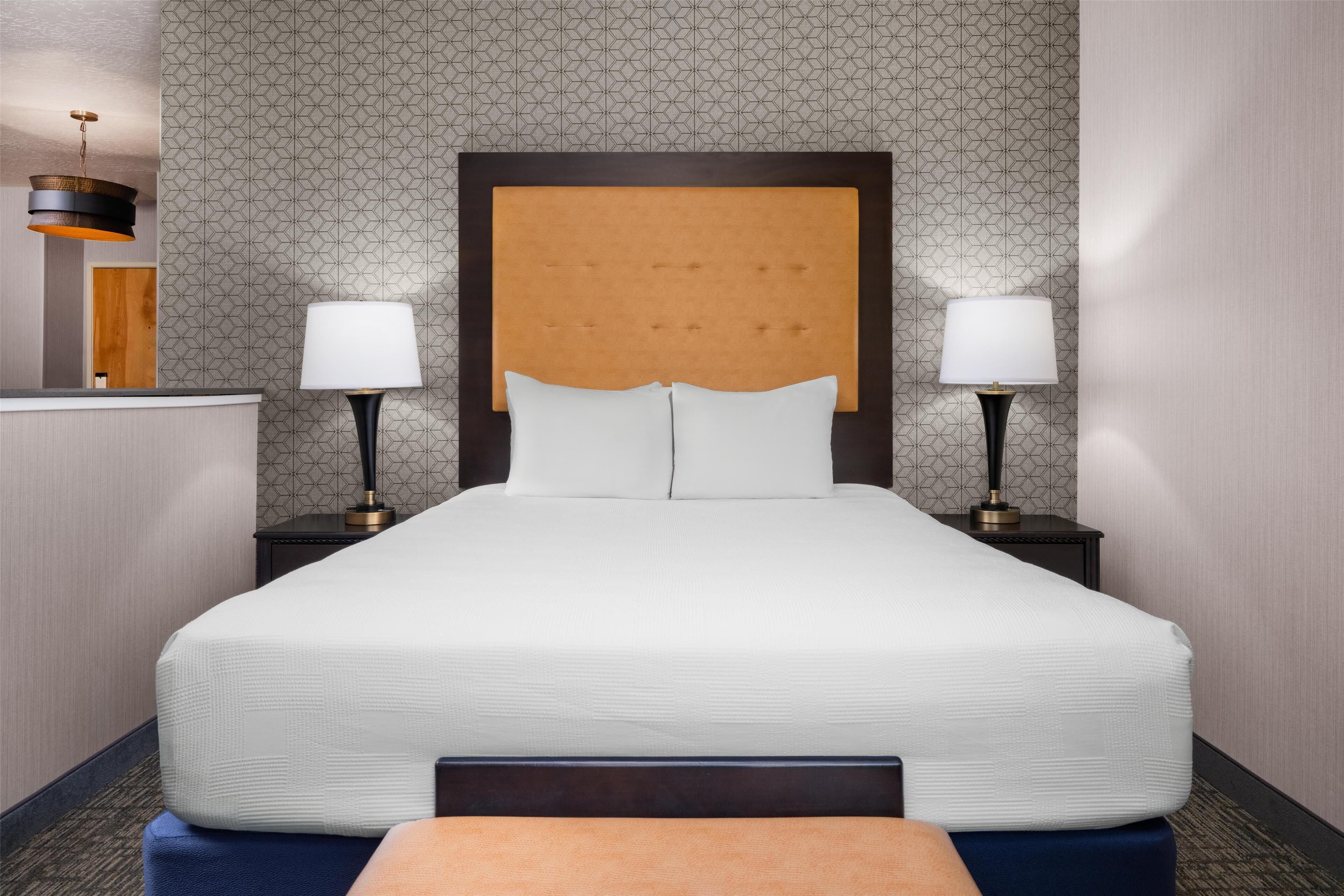 A hotel room with a large bed, two bedside tables with lamps, a headboard, and a patterned wallpaper.
