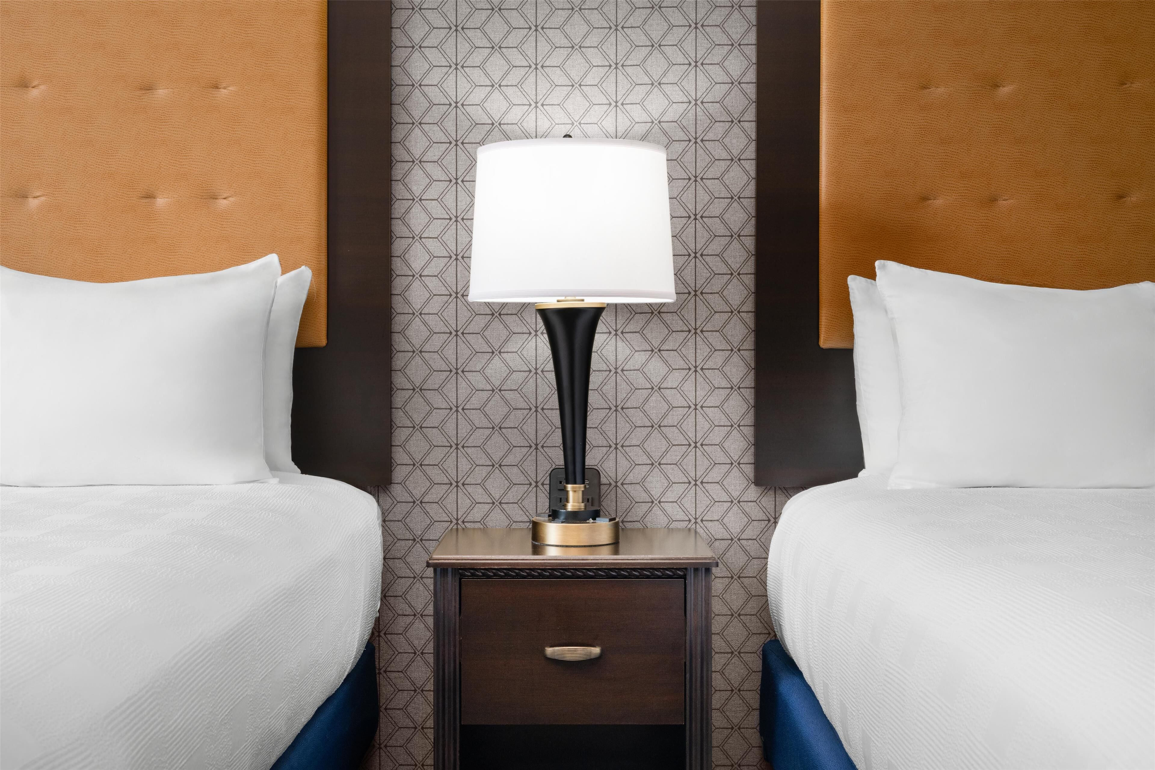A hotel room with two beds, a nightstand with a lamp, and a patterned wallpaper.