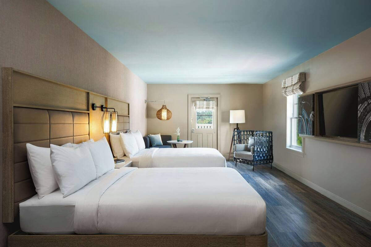 A hotel room with two beds, a small table, a chair, a TV, and a window with a view.