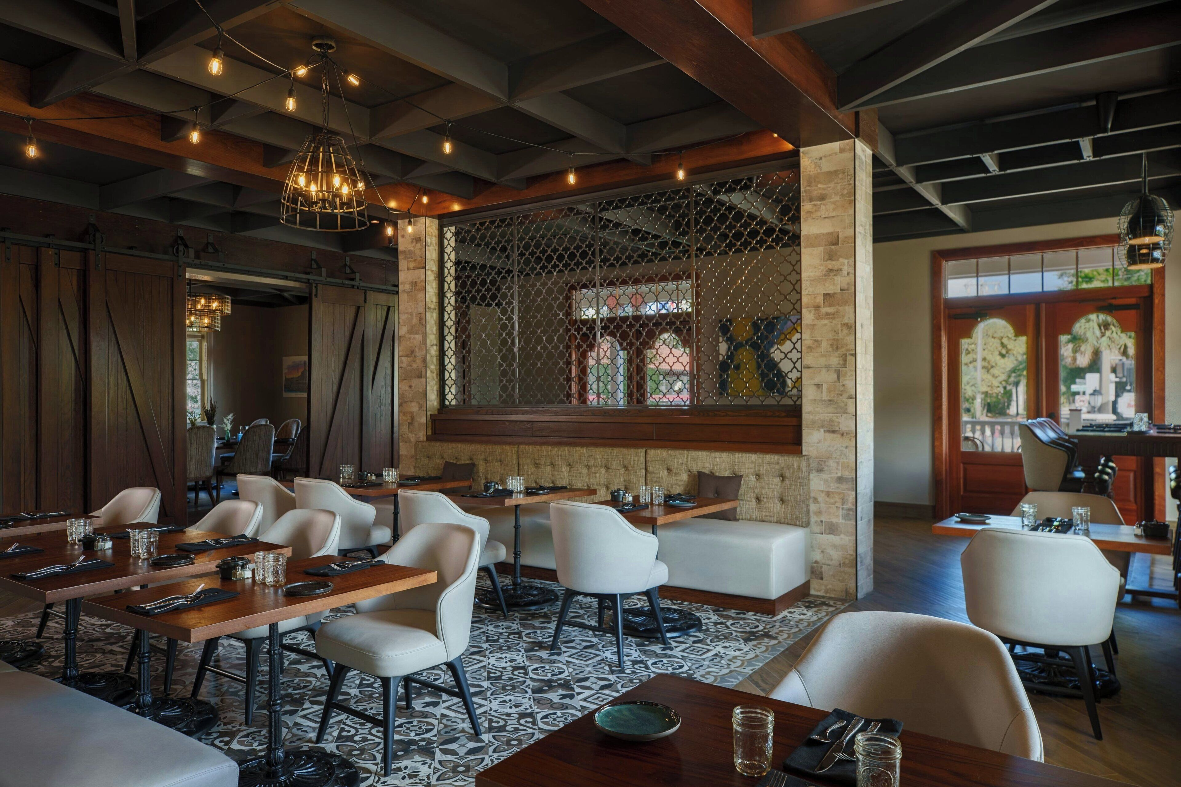 A restaurant with wooden tables and white chairs, a patterned rug, and a large window with a view.