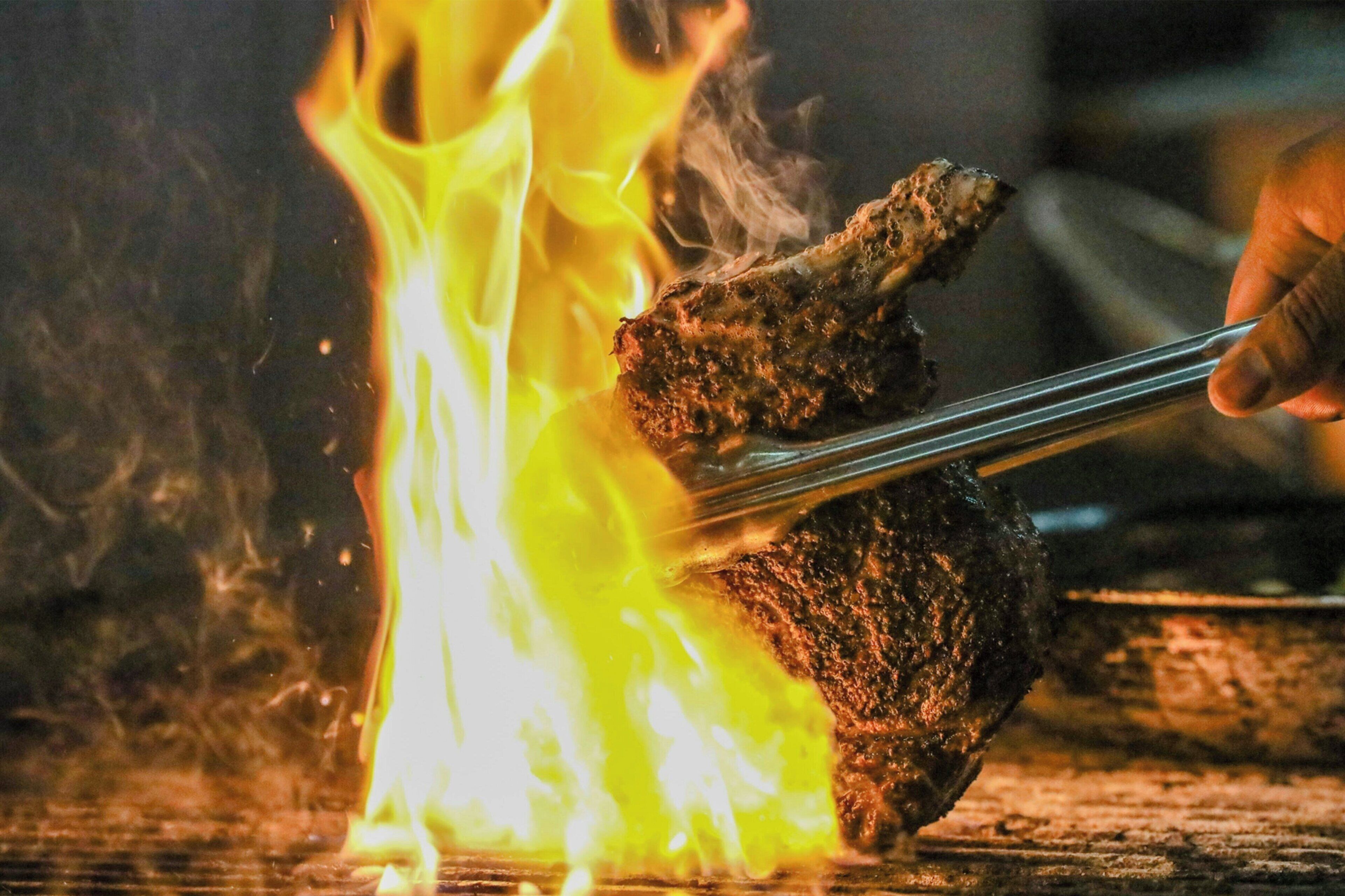 A grilled meat being held with tongs over a fire.