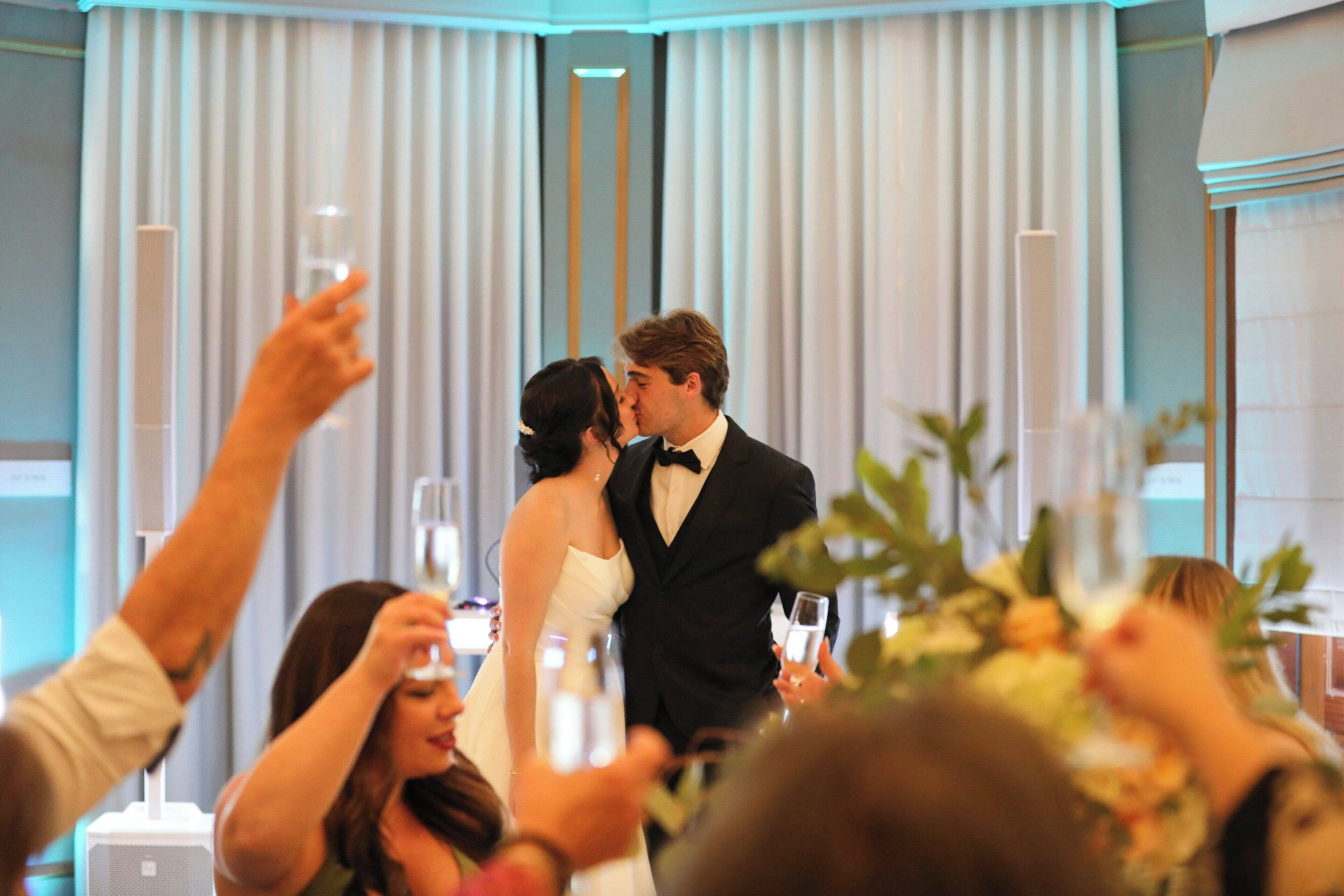 Une réception de mariage où l’on voit un couple s’embrasser, des invités tenant des verres et une composition florale au premier plan.