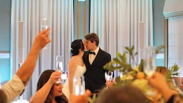 Une réception de mariage où l’on voit un couple s’embrasser, des invités tenant des verres et une composition florale au premier plan.