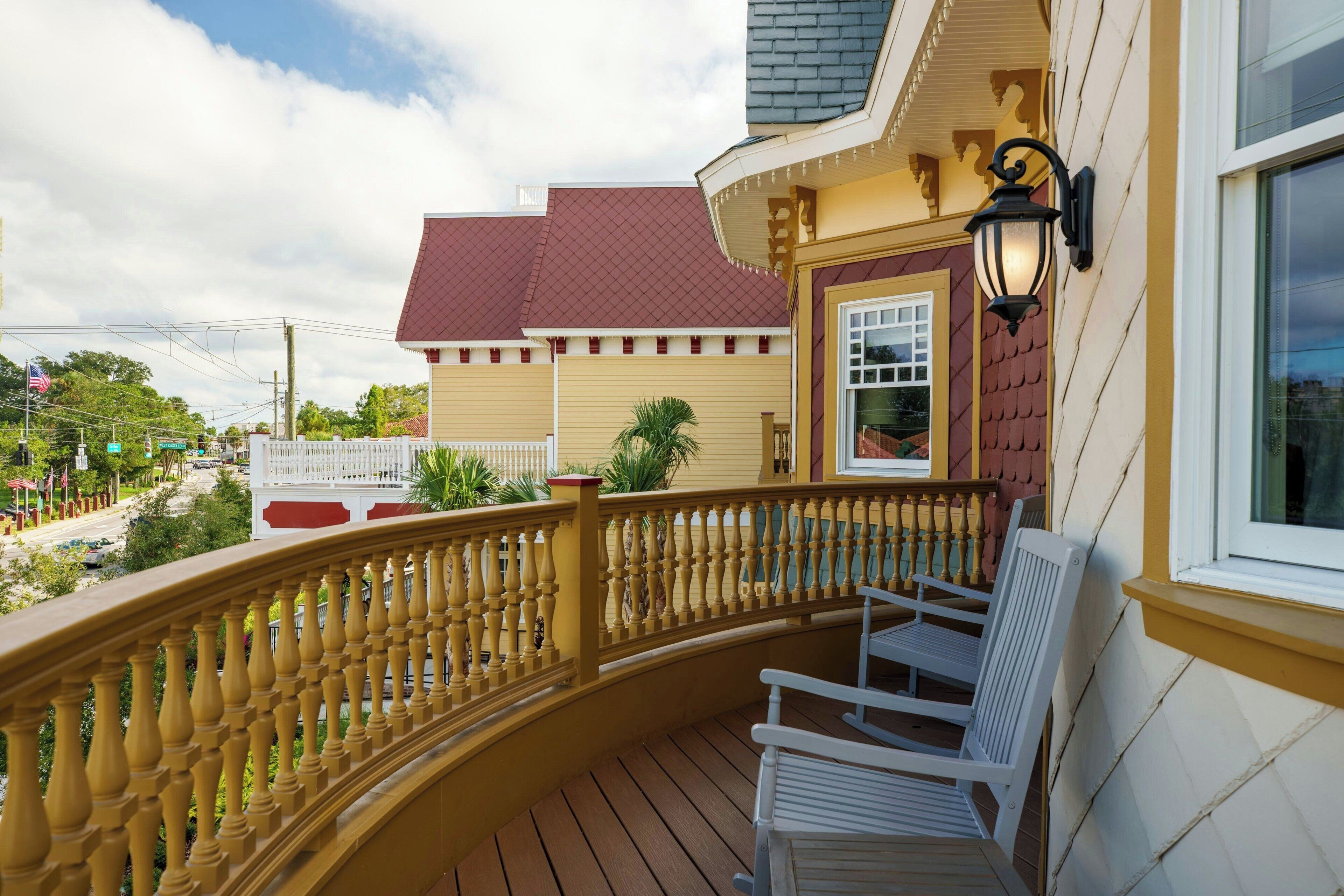 A wooden deck with a railing, white chairs, and a building with a red roof and beige walls.