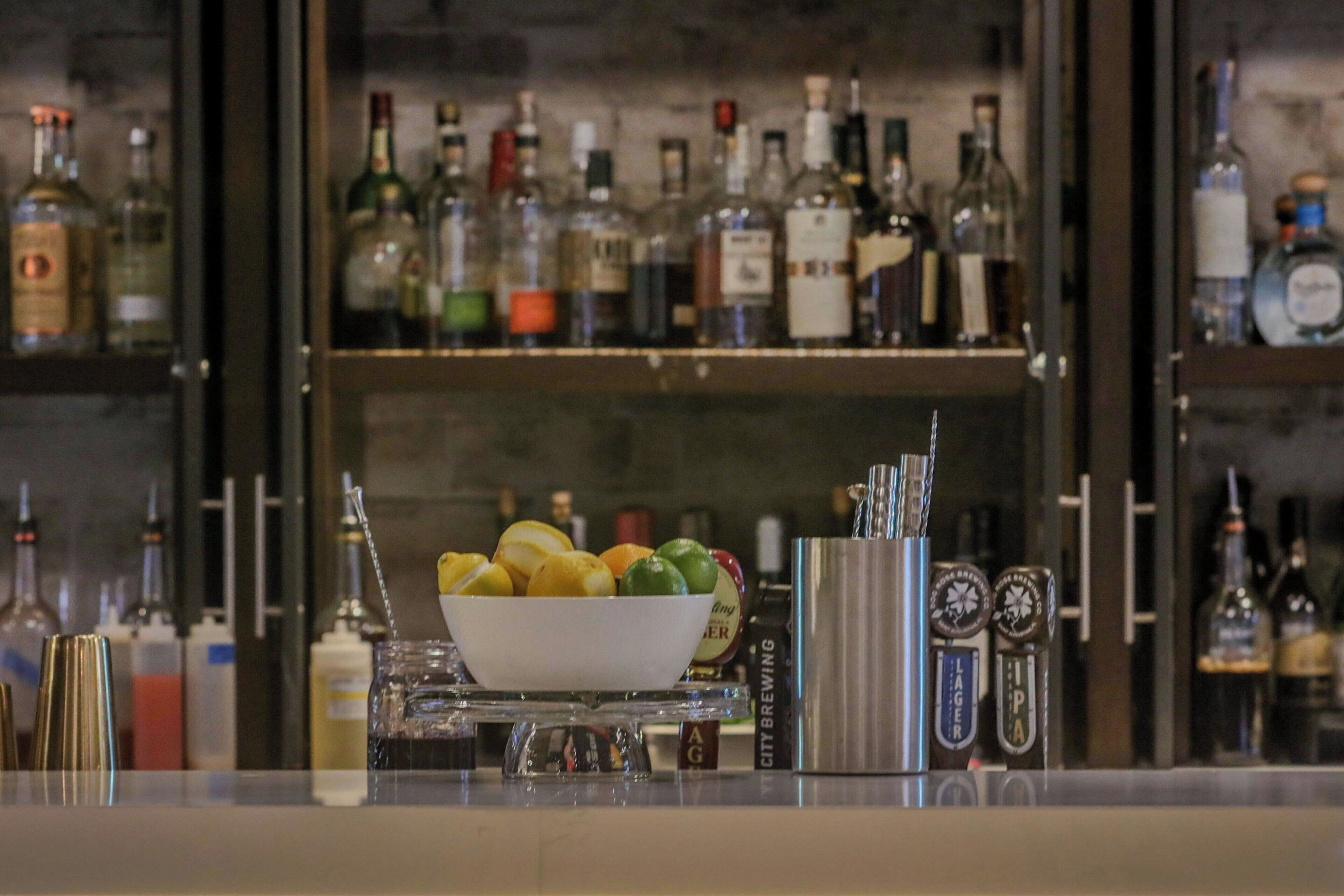 Una zona de bar con varias botellas de licor y un frutero sobre un pedestal.