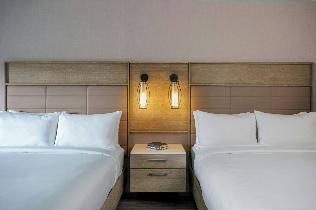 A hotel room with two beds, a wooden headboard, bedside lamps, and a small bedside table with a book.