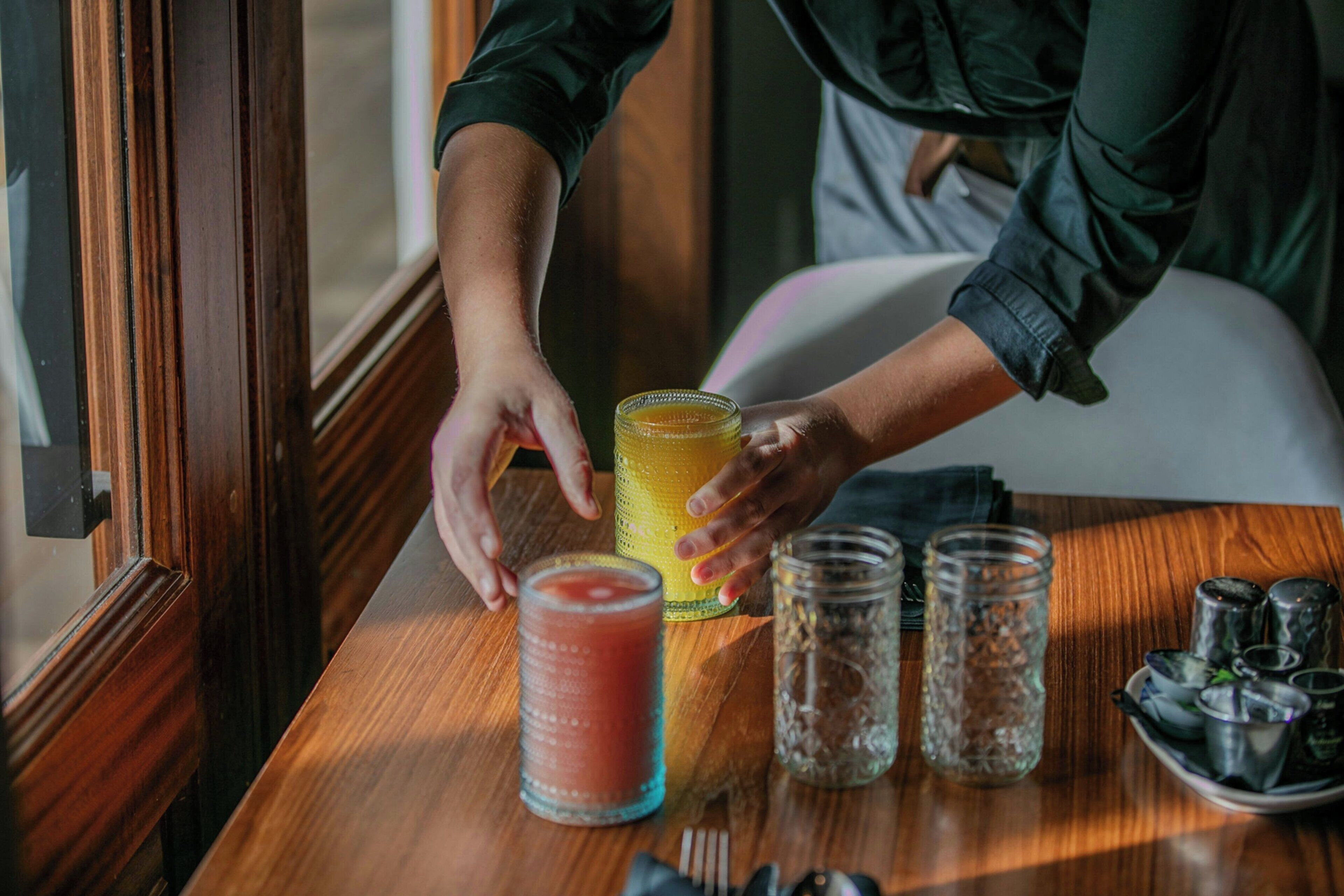 Una mesa de madera con un vaso de bebida roja, una bebida amarilla y varios vasos y botellas.