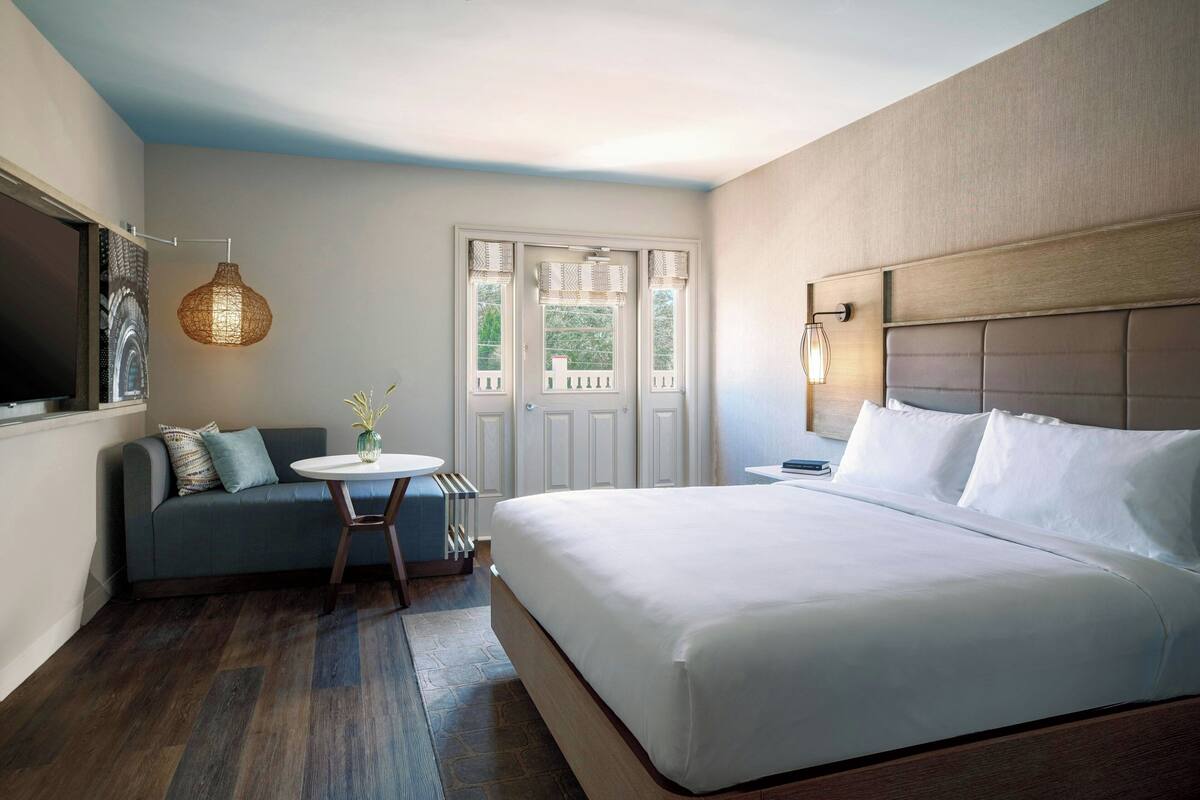 A modern hotel room with a large bed, a sofa, a small round table, and a view of the outdoors through a door with windows.