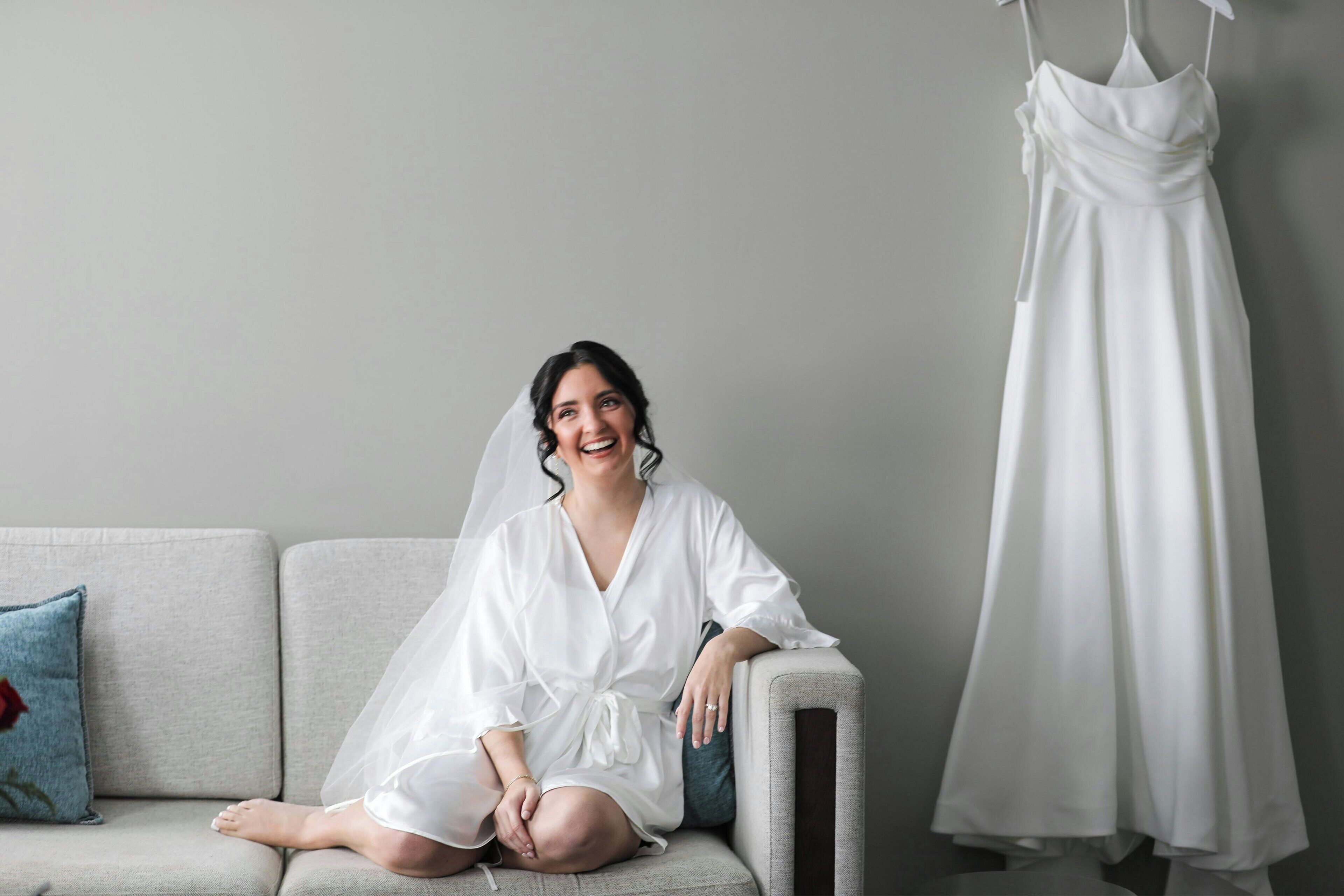 A person sitting on a sofa with a white dress and veil, a dress hanging on the wall, and a blue pillow on the sofa.
