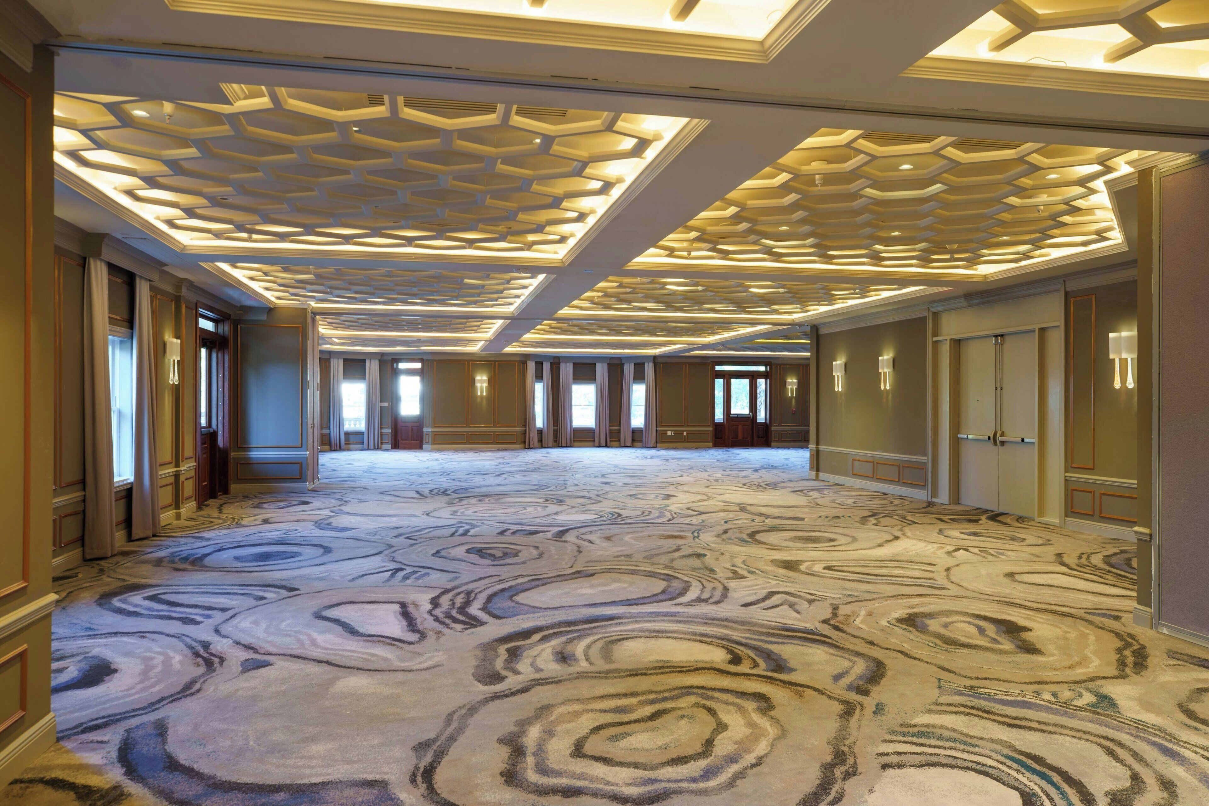 Un hall d’hôtel spacieux, doté d’un tapis à motifs, d’un plafond au design géométrique et d’un long couloir bordé de portes.