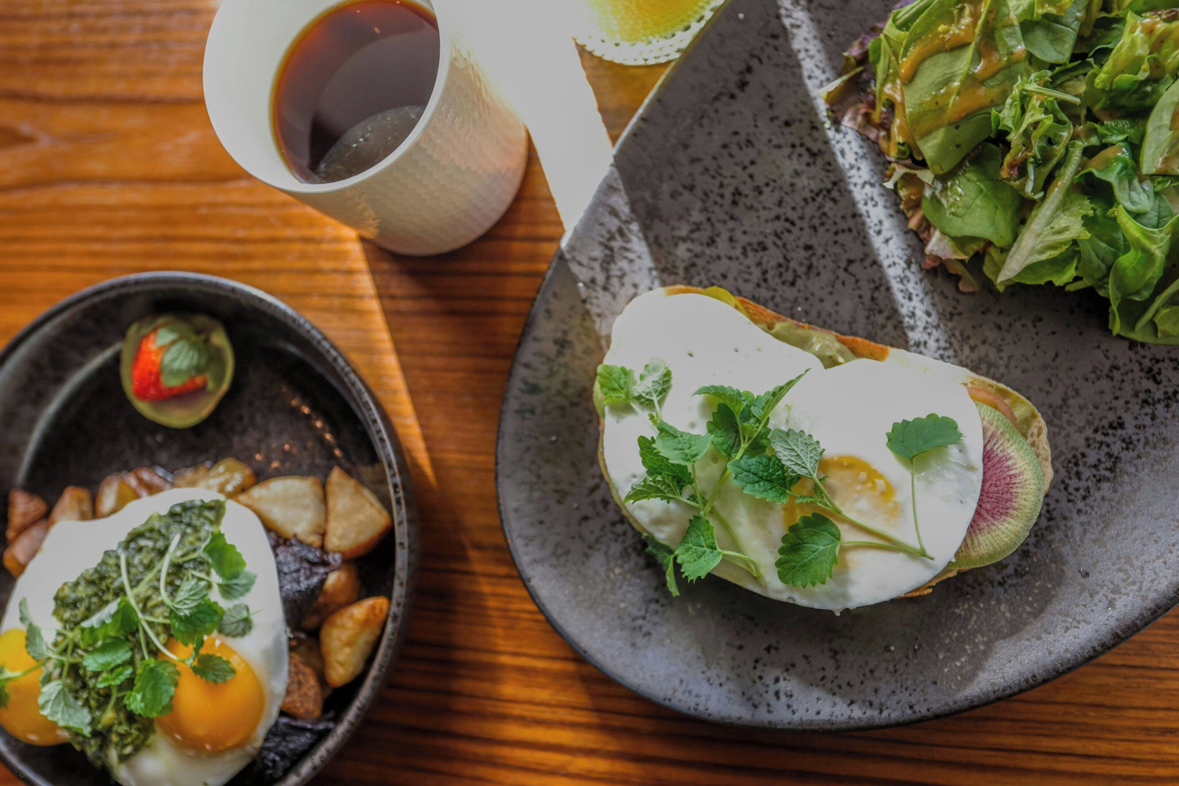 A dining table with a variety of dishes including a plate with a white sauce and greens, a bowl with a green sauce and egg, and a cup of tea.