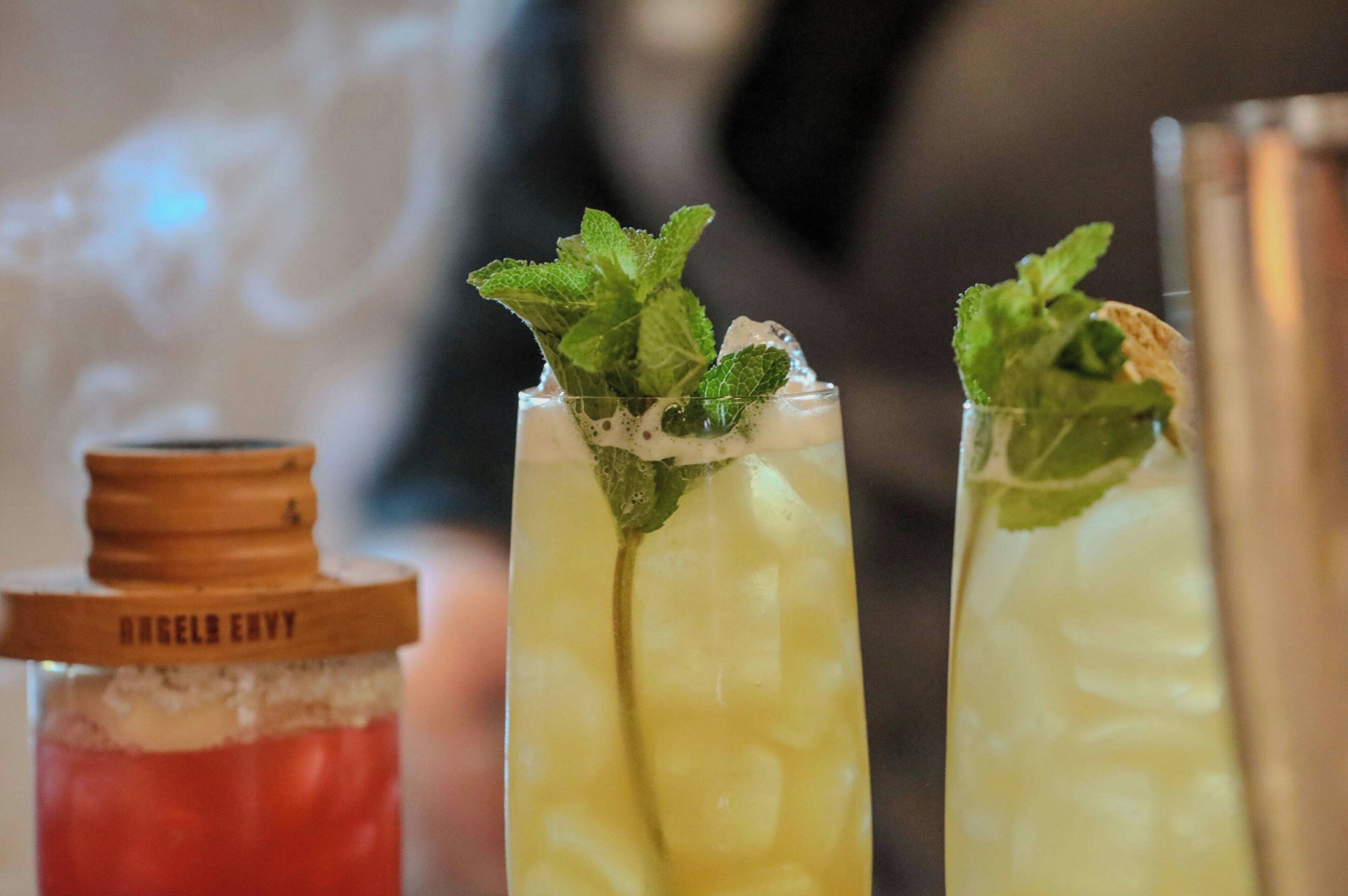 Three glasses of a yellow beverage with mint leaves, a bottle with a wooden lid, and a metal shaker.