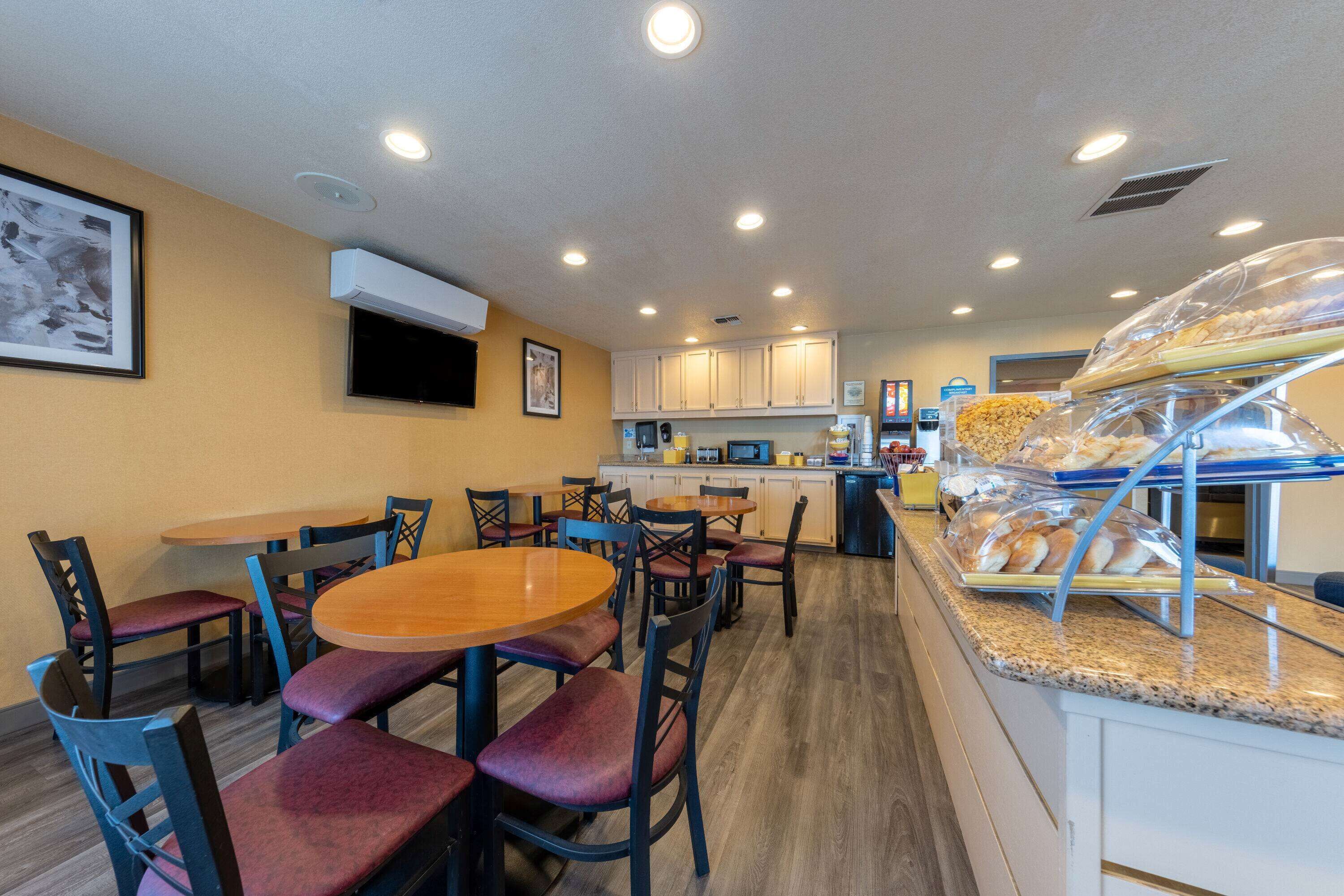 A restaurant with round tables and chairs, a kitchen area, and a display case with bread.