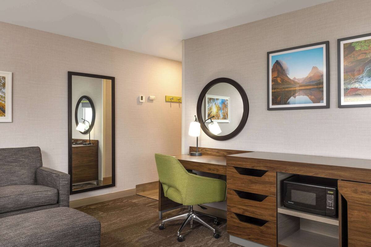 A hotel room with a grey sofa, a green office chair, a wooden desk with a built-in microwave, and two framed pictures on the wall.