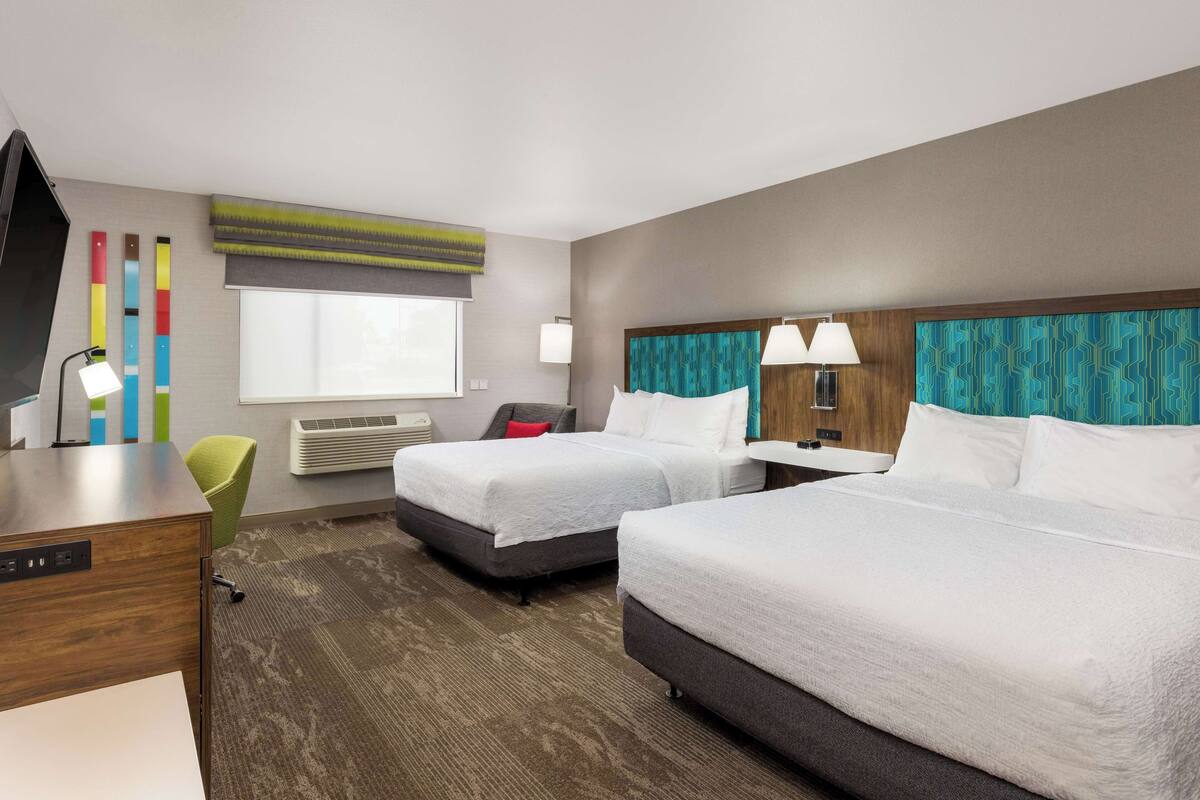A hotel room with two beds, a desk, a chair, a TV, and a window with blinds.