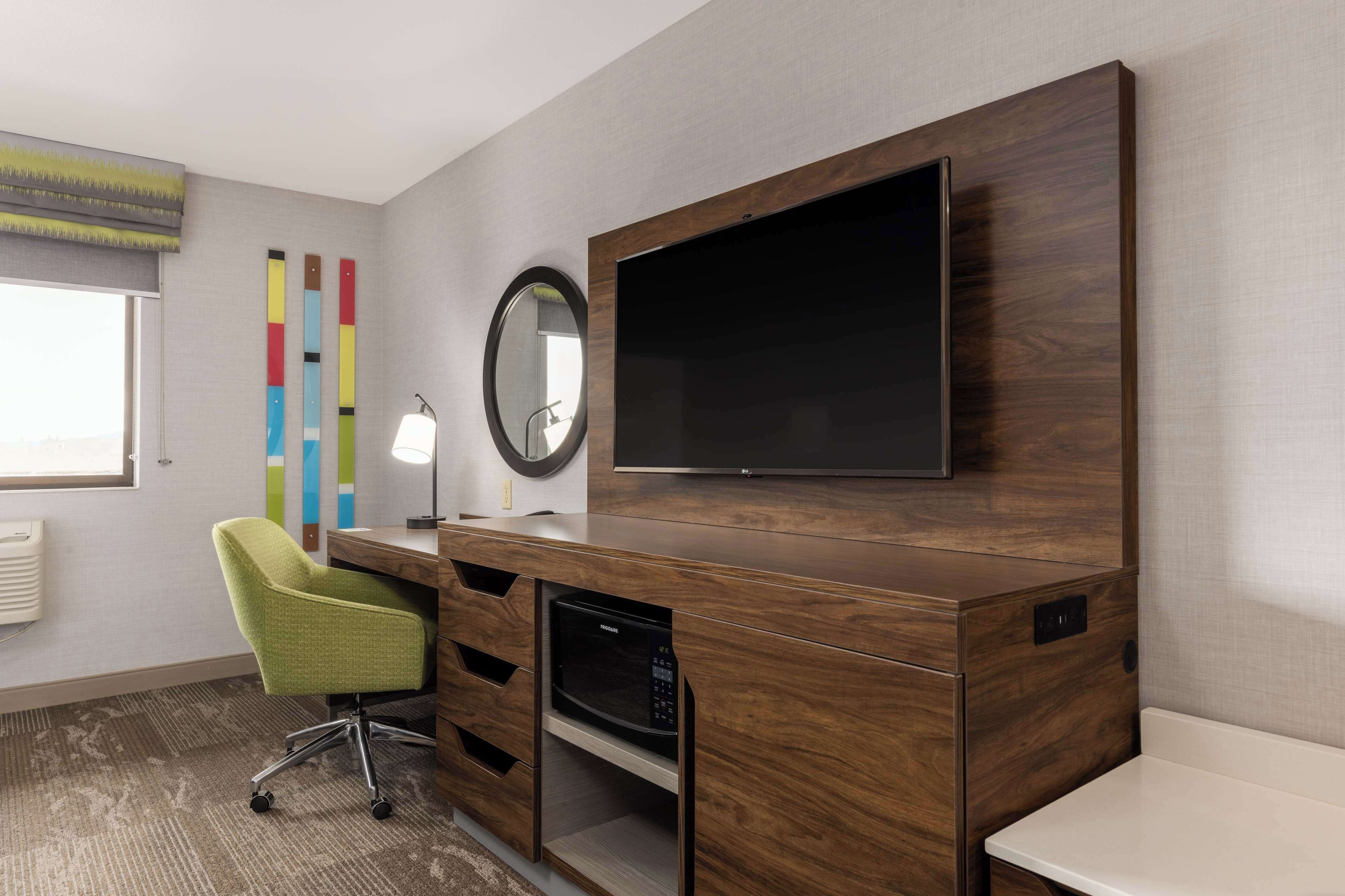Una habitación de hotel moderna con un escritorio de madera, un televisor de pantalla plana, una silla verde, una lámpara de escritorio y un espejo grande.