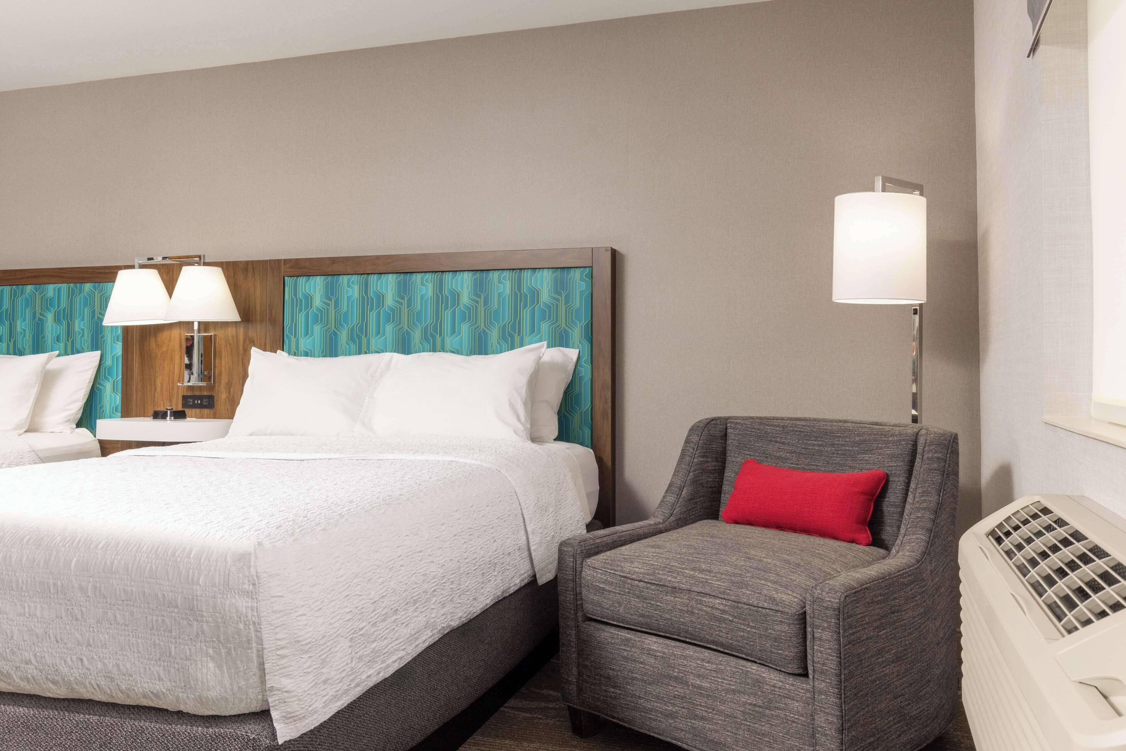 Una habitación de hotel con una cama, dos lámparas de noche, una silla con un cojín rojo y un calentador blanco.