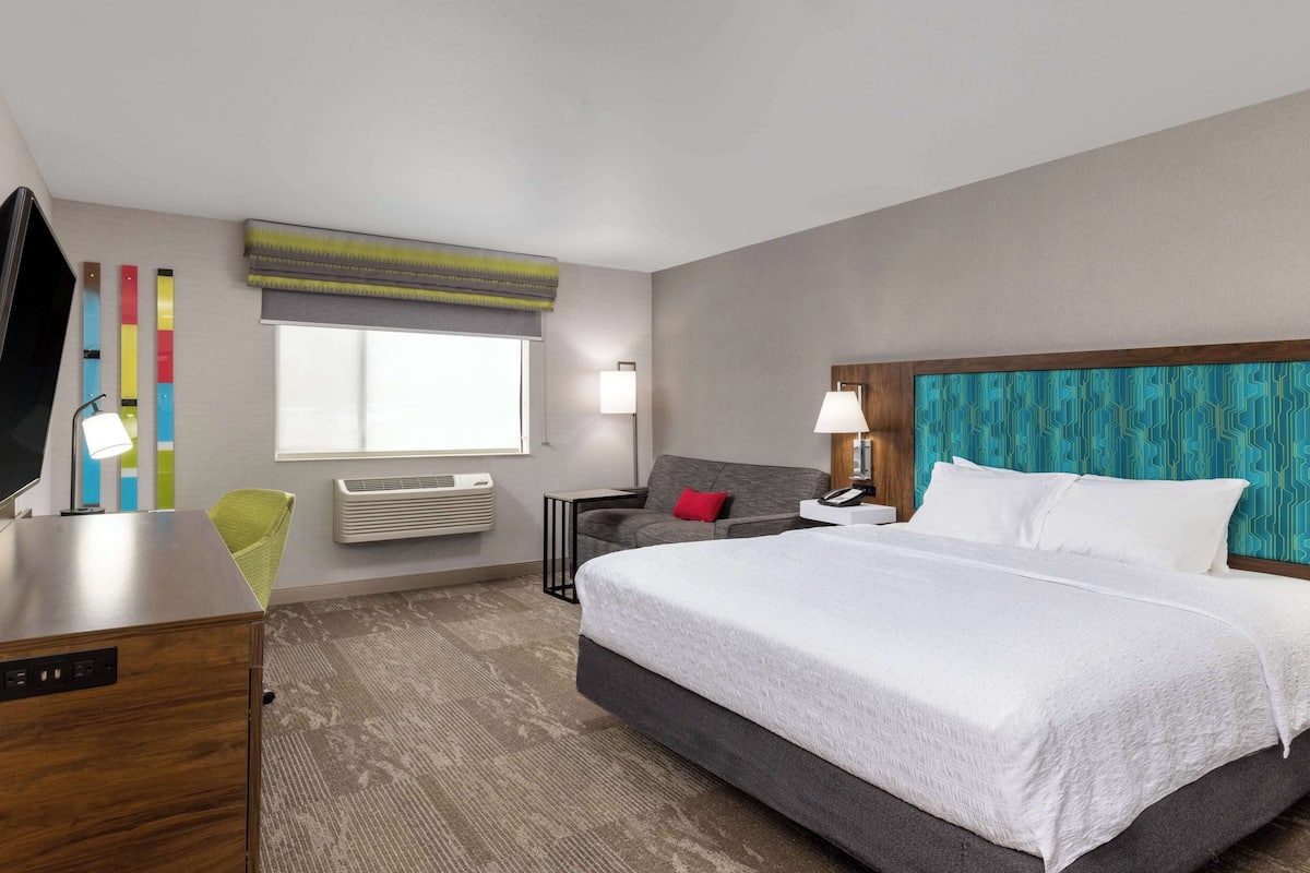 A hotel room with a large bed, a desk, a chair, a TV, a window with blinds, and a wall with a patterned design.