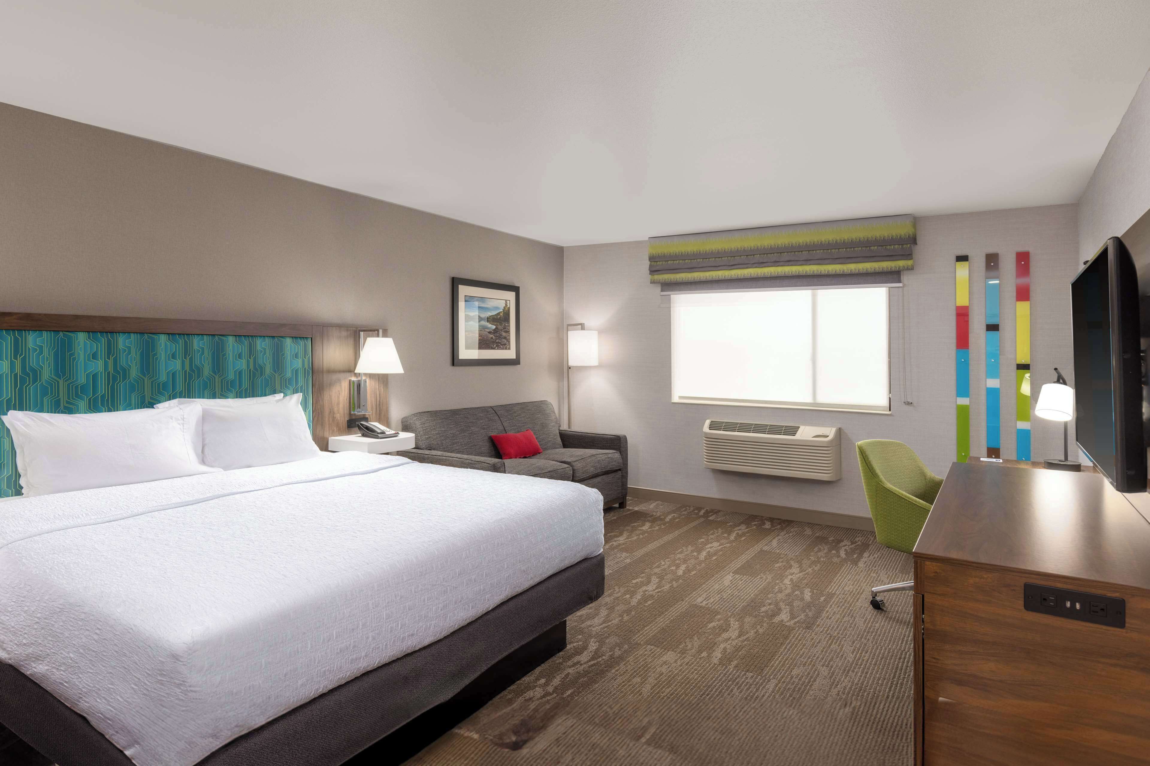 A hotel room with a large bed, a desk, a chair, a sofa, a TV, and a window with blinds.