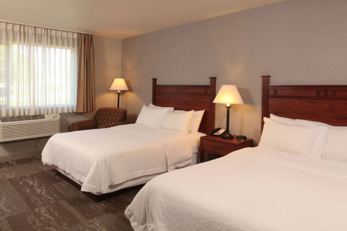 A hotel room with two beds, a chair, bedside lamps, an air conditioner, and a window with curtains.
