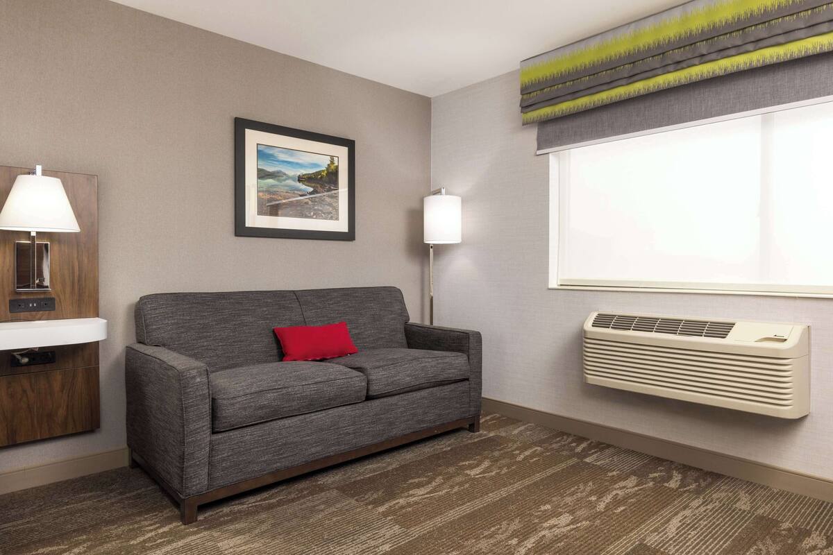 A room with a grey sofa, a red pillow, a framed picture, a wall-mounted air conditioner, and a window with blinds.