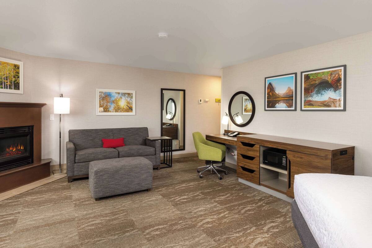 A hotel room with a fireplace, grey sofa, ottoman, desk with chair, and artwork on the walls.